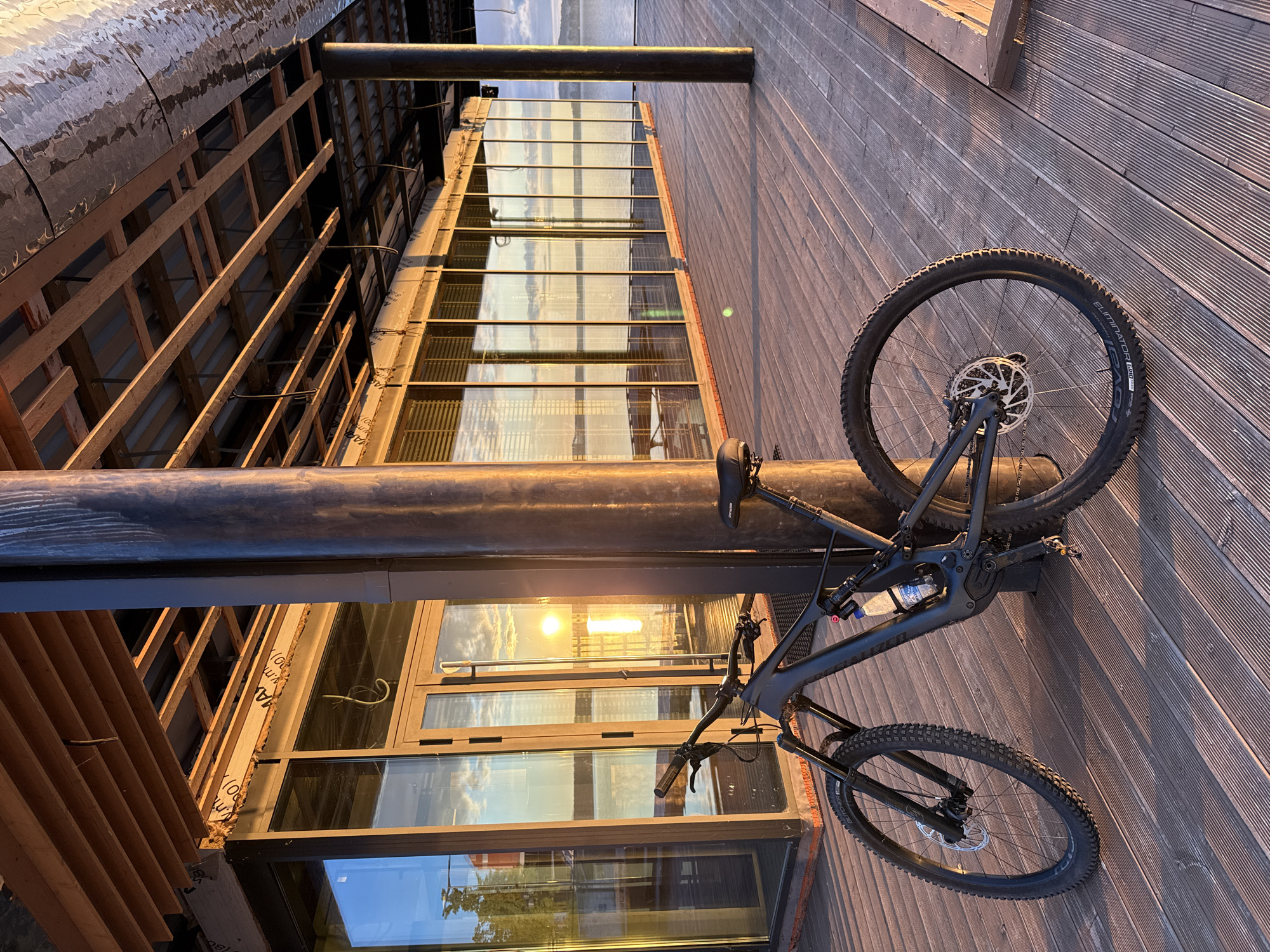 The image, correctly oriented, displays a black full-suspension mountain bike parked on a wooden deck or boardwalk, leaning against a dark metal pillar. The scene is illuminated by the warm, golden light of the setting sun, which creates prominent reflections in the large glass windows of an adjacent modern building. Beyond the building, a body of water is visible, also reflecting the sky.
The side of the building closest to the viewer features an exposed architectural element or is under construction, revealing a framework of horizontal wooden slats and beams over a dark, textured weather-resistant material. Faint text like "VAPOR" and "AEROTEC" is visible on some of these building materials.
The mountain bike is a prominent subject, featuring "ELIMINATOR GRID TRAIL" and "TL READY" branding on its tires, consistent with Specialized models, with "SPECIALIZED" also faintly visible on the downtube. A water bottle, marked "AGUA", is secured in the bike's frame. The bike is equipped with disc brakes.
The overall atmosphere suggests late afternoon or early evening, characterized by soft light and long shadows. The location is Votkinsk, Russia.