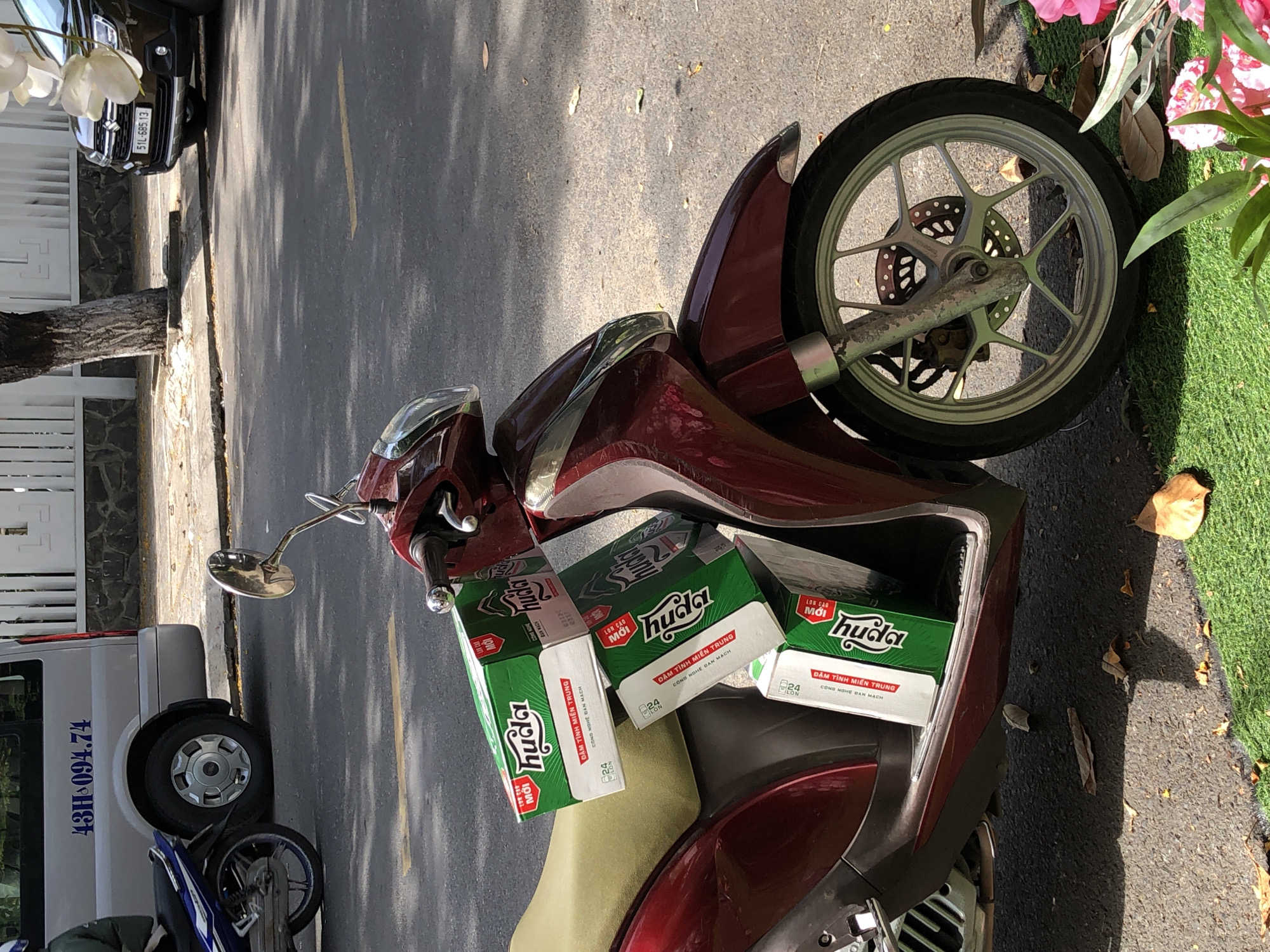 In Da Nang, Vietnam, a maroon scooter is parked on a paved street. The scooter is loaded with stacked green boxes of what appears to be beer, with the brand name "Huda" visible. The boxes are positioned on the seat and over the handlebars. In the background, a silver van with license plate "43H-094.74" is parked. To the right of the scooter, a dark brown car with license plate "51L-685.13" is also parked. The street is lined with a white fence and a tree. Some dry leaves are scattered on the ground. The scene appears to be a typical urban setting.