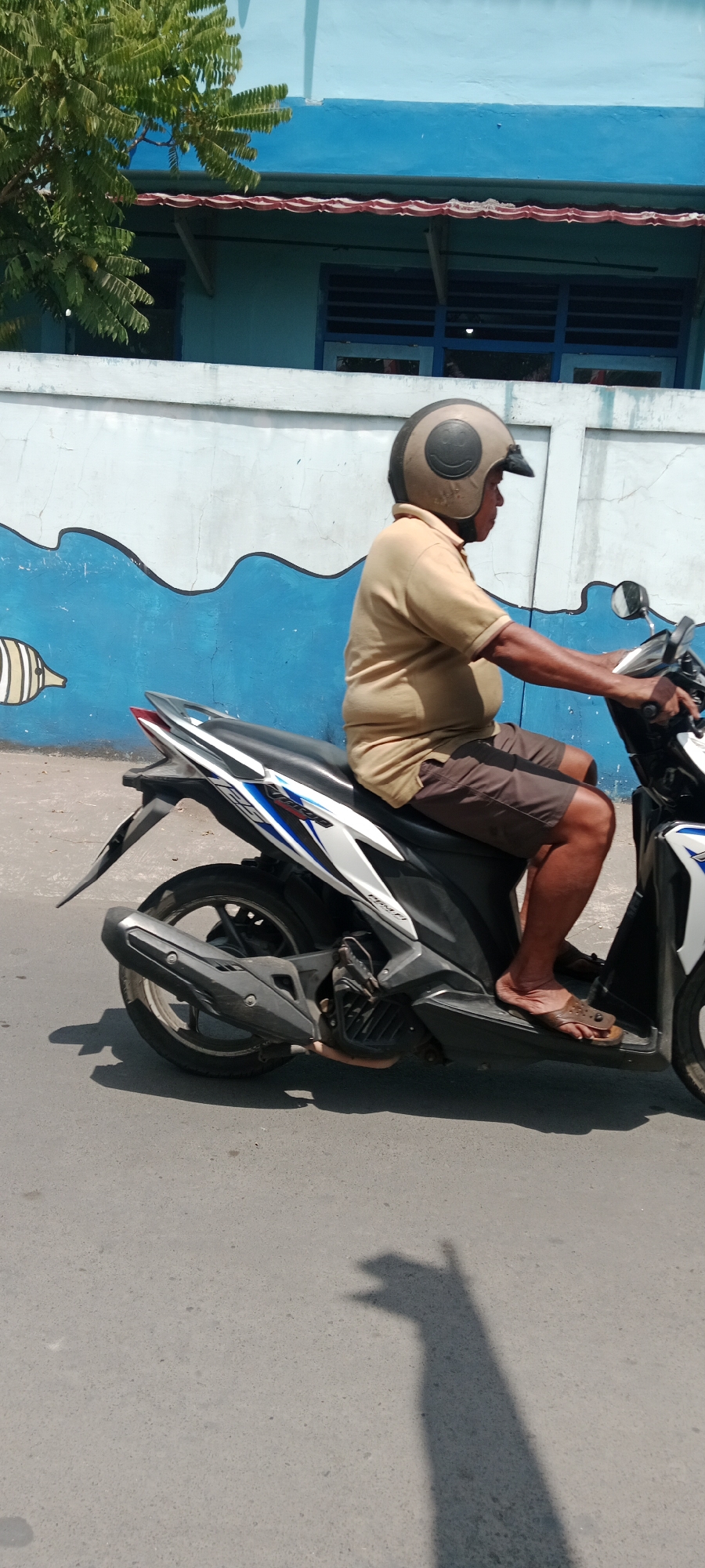 An adult male is riding a scooter on a street in Purwomartani, Indonesia. He is wearing a light brown polo shirt, brown shorts, and brown flip-flops. He has a beige helmet with a smiley face design on it. The scooter is white, black, and blue. The background consists of a blue wall with a white wave mural and a green tree. The sun casts a shadow of the scooter and rider on the asphalt road.
