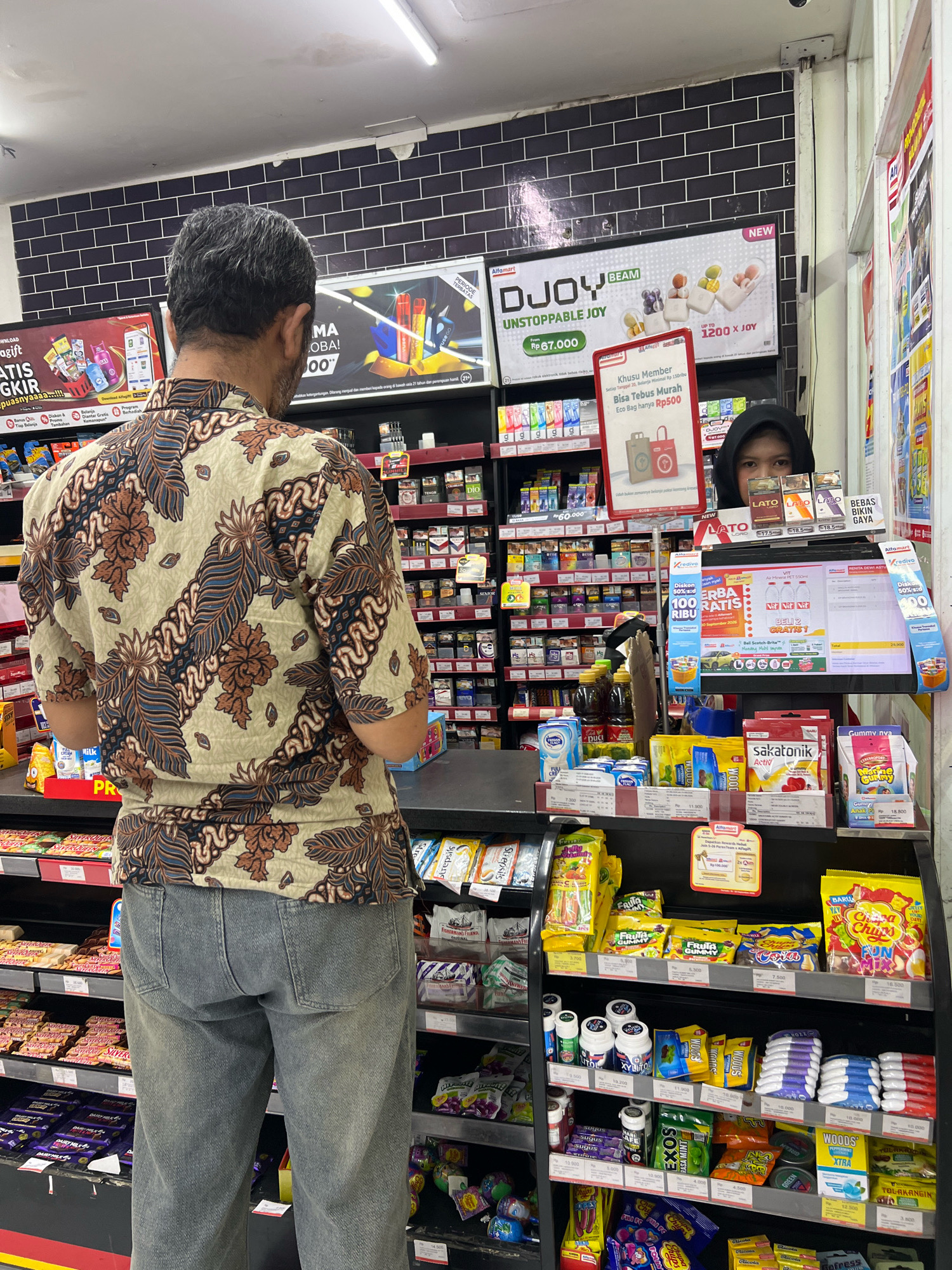 In a brightly lit convenience store in Tlogoadi, Indonesia, a man with graying hair, wearing a batik shirt and denim jeans, stands with his back to the camera, facing a counter laden with various products. Behind the counter, a young woman wearing a black hijab is partially visible, her eyes looking directly at the camera. The shelves behind them are stocked with an assortment of packaged goods, including snacks, drinks, and personal care items. Promotional displays are visible on the walls and counter, featuring advertisements for various brands and offers. The overall scene suggests a typical retail environment within the city.