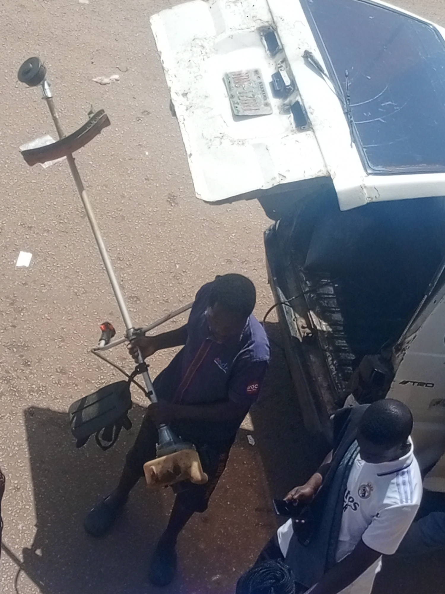 Two men are shown in a scene that appears to be outdoors, with a gritty, asphalt-like ground. In the foreground, a person wearing a dark blue shirt with a logo that reads "AGC" and dark trousers is holding a weed trimmer. The weed trimmer has a long shaft and a cutting head at the bottom. The man is also wearing dark shoes and appears to be in the process of using or adjusting the equipment.
Behind him, a white vehicle is visible, partially overturned or damaged. The vehicle's tailgate is open, and the interior is dark. A license plate is visible on the back of the vehicle, though the text is not clearly legible. The car's rear window is dark and appears to be cracked.
To the right of the man with the weed trimmer, another man is partially visible. He is wearing a white t-shirt with blue stripes on the sleeves and a crest that resembles a sports team logo, possibly Real Madrid. He has a dark scarf or cloth draped over his shoulders and is holding a dark object, possibly a phone, in his hand.
The lighting suggests it is daytime, likely with sunny weather given the shadows. The overall scene suggests a task related to yard work or maintenance, possibly in a residential or open area in Bukuru, Nigeria. There are no strong indicators of specific emotions, but the focus of the individuals on their tasks is evident.