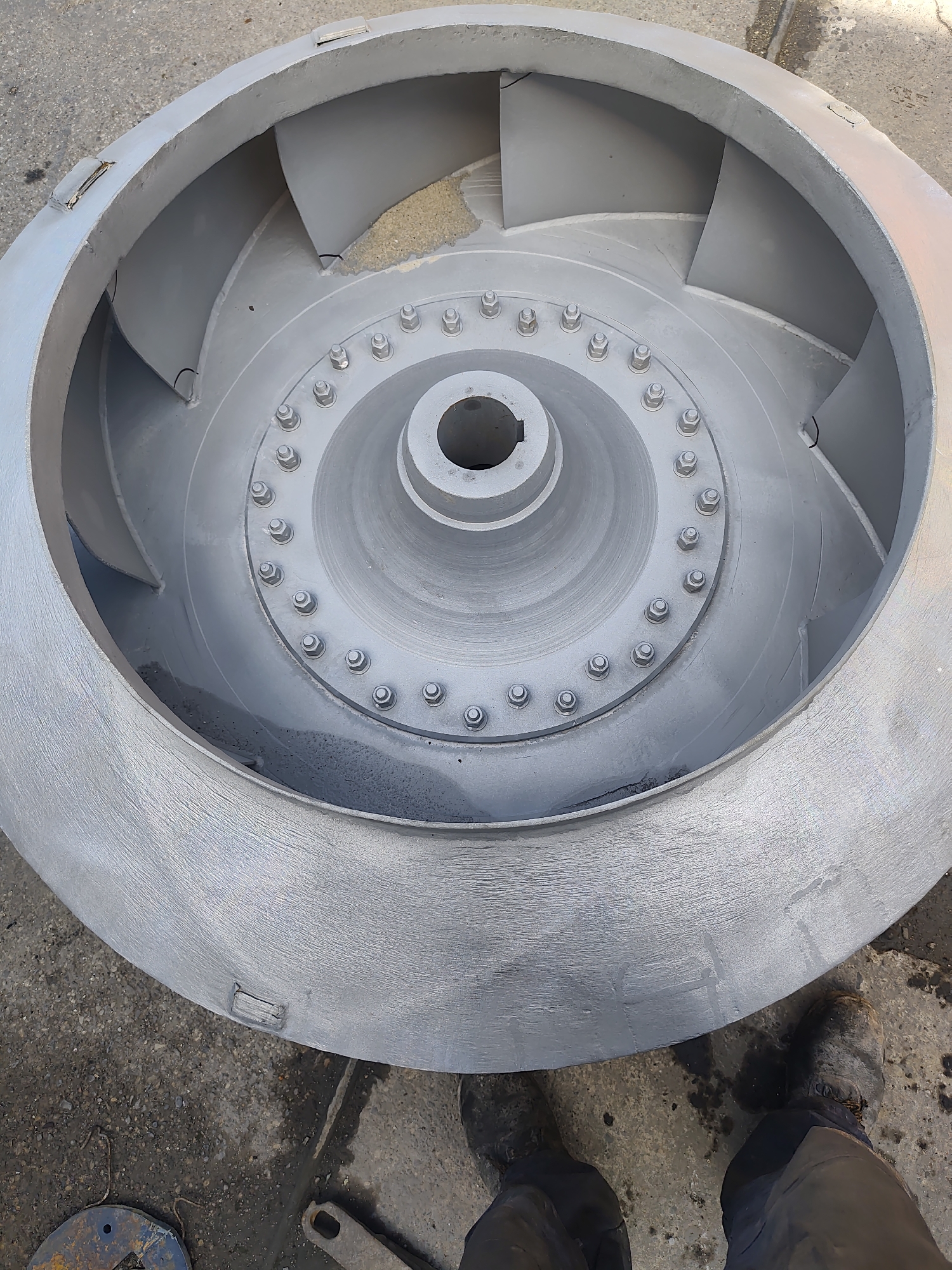 An overhead, slightly angled view shows a large, industrial fan impeller, likely made of metal and painted a matte gray, lying flat on a concrete surface. The impeller is circular, with a central hub featuring a square opening for a shaft. Radiating outward from the hub are multiple curved vanes. The outer rim of the impeller is thick and robust, with several square-shaped brackets or mounting points visible along its exterior.
The impeller is surrounded by a rough, gray concrete surface that appears to be outdoors, possibly in a service area or workshop. The ground is slightly textured and shows some darker stains and small debris. In the bottom right corner of the image, the legs and feet of a person wearing dark pants and dirty work boots are visible, suggesting the impeller is either being worked on or has recently been moved. The scene appears to be located in an industrial setting, and while the prompt requests the city of Police, Poland, there are no specific visual cues within the image to confirm this location.