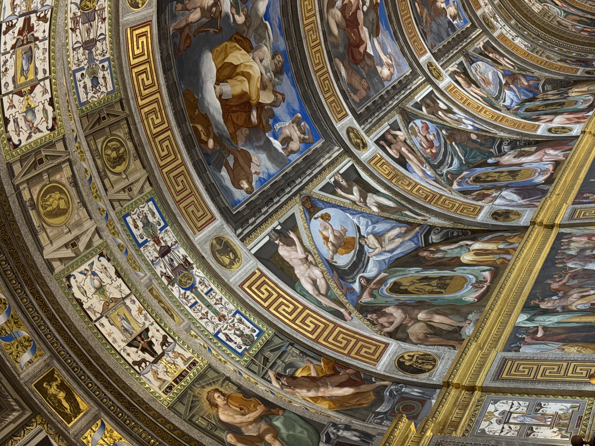 This image depicts the opulent ceiling frescoes of the Biblioteca de el Escorial in San Lorenzo de El Escorial, Spain. The artwork covers the vaulted ceiling of a grand hall, showcasing intricate mythological and allegorical scenes rendered in vibrant colors. The frescoes are divided into rectangular and arched sections, separated by gilded borders and decorative friezes featuring Greek key patterns.
The artwork features numerous human figures, predominantly male, depicted in various poses and engaged in different activities. Many figures are nude or semi-nude, rendered with idealized anatomy. Some figures are shown playing musical instruments, such as a lyre or a horn. Others are depicted in dynamic poses, suggesting movement or dramatic action. Celestial bodies, such as clouds and sky, are visible in some of the painted sections, adding a sense of depth and grandeur.
The overall impression is one of immense artistic skill and a rich narrative tapestry. The architectural elements of the vaulting are emphasized by the painted decorations, creating a cohesive and immersive visual experience. The lighting appears to be natural, possibly from unseen windows or skylights, highlighting the textures and colors of the frescoes. The environment is clearly an indoor, highly decorated space, likely a library or a grand hall within a significant historical building.