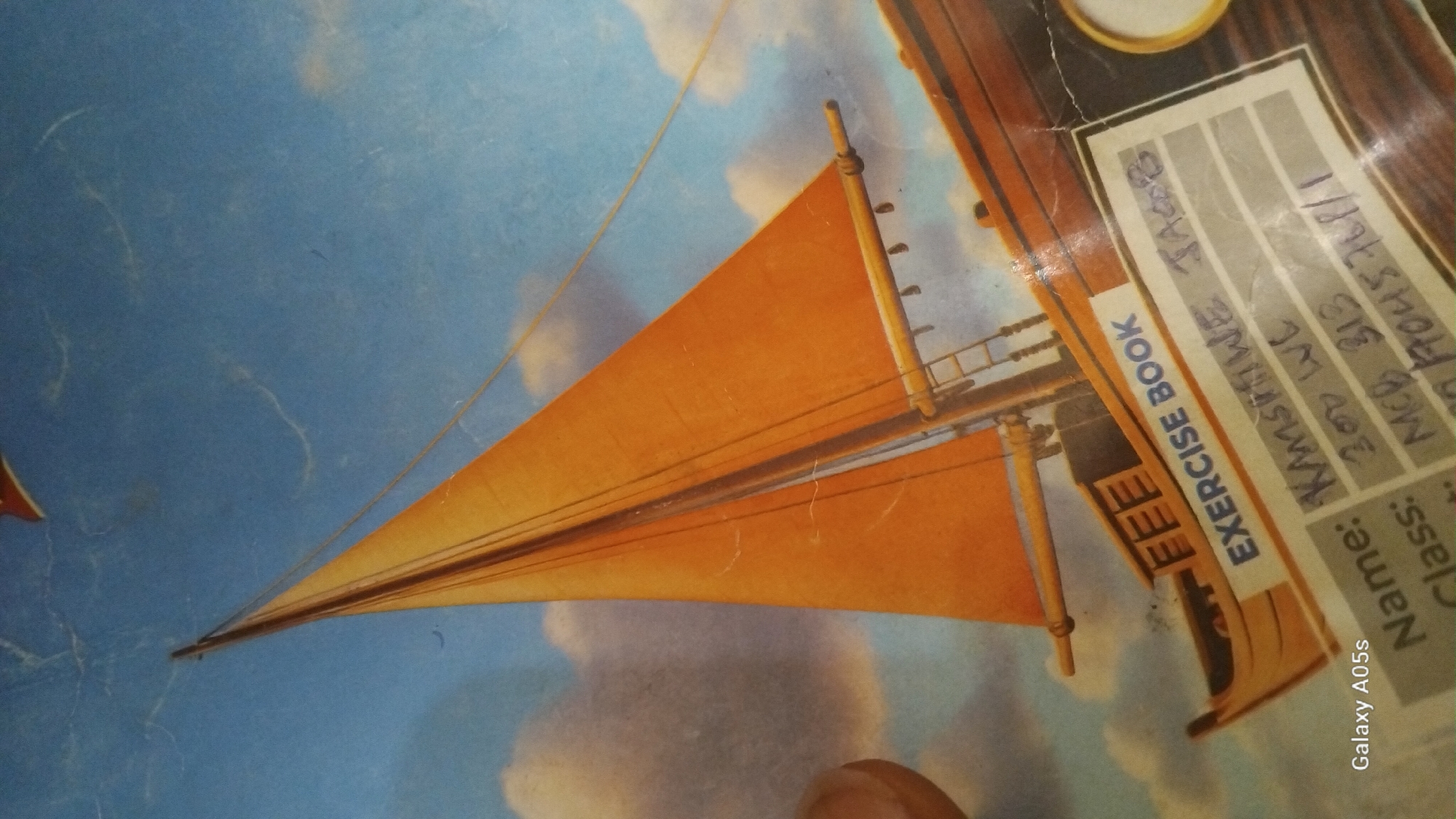 The image displays a notebook cover featuring a painting of a sailboat with orange sails against a blue sky with clouds. Text on the notebook is partially visible, including "EXERCISE BOOK" and handwritten entries for "Name:", "Class:", and a number. The bottom left corner shows the text "Galaxy A05s". The notebook is slightly worn, with visible scuffs and scratches on the cover. This notebook is likely located in Yali, Nigeria, as indicated by the user's prompt. The scene is static and does not depict any active events or people in the immediate foreground.