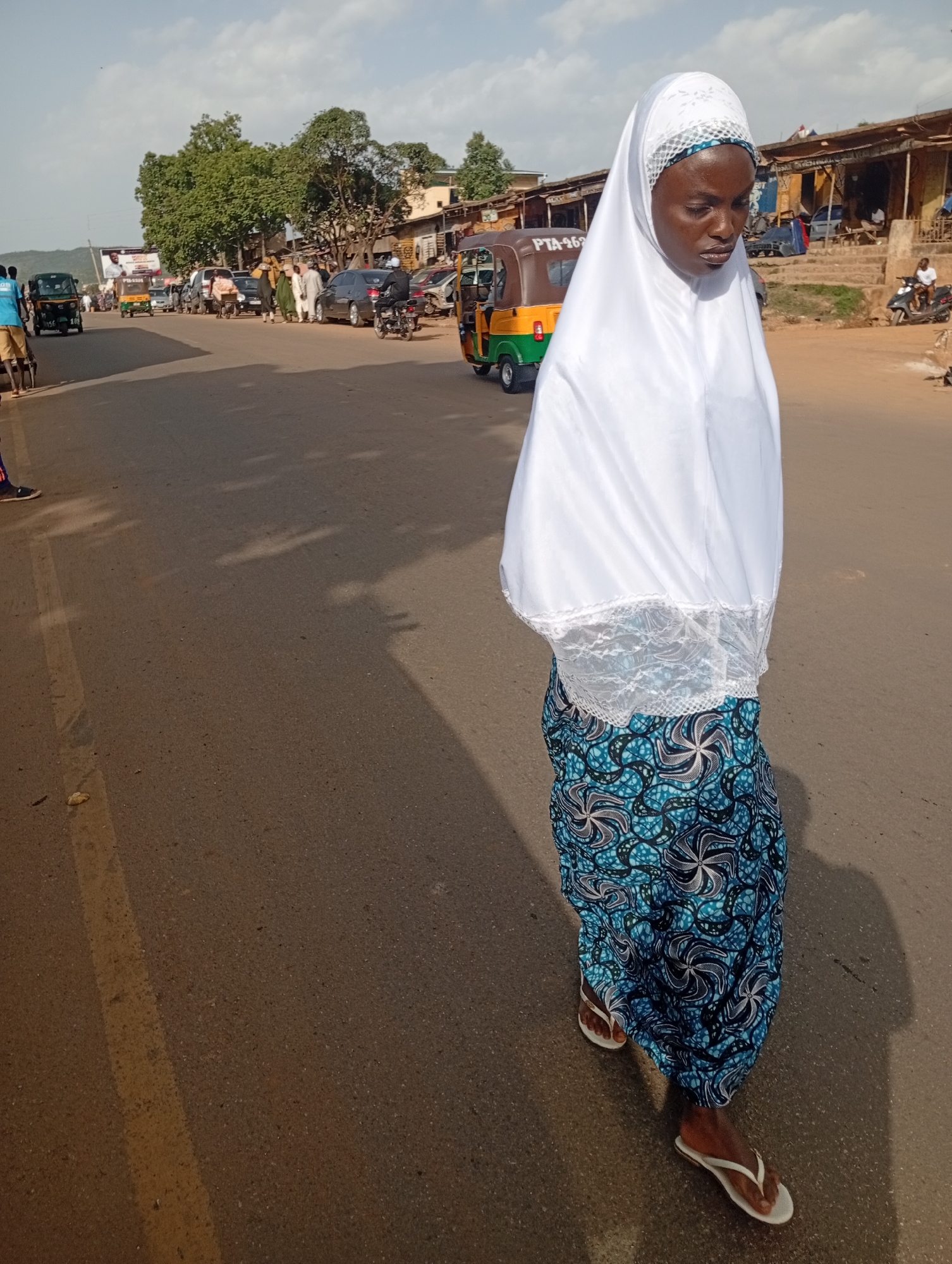 A young Nigerian woman wearing a white hijab and a patterned blue and black skirt walks on a paved road in Rafin Par, Nigeria. She is wearing white flip-flops. The road is lined with shops and buildings on the right side of the image. Several vehicles, including a tricycle and motorbikes, are visible on the road. The sky is partly cloudy. The woman is walking towards the right side of the frame.