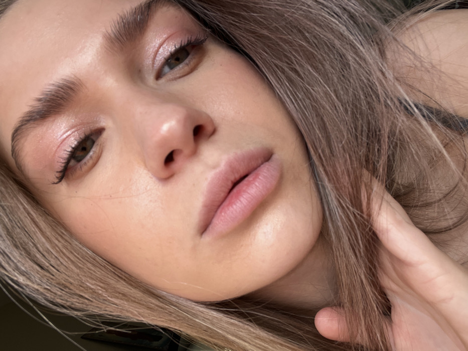 The image presents a close-up, angled view of a young adult female's face and upper torso, taken in a selfie style. The primary subject is a female with fair skin, light brown or blonde hair, and brown eyes. Her eyes are open, and she is looking directly or slightly away from the camera. She has applied light makeup, including pink eyeshadow on her eyelids, black winged eyeliner, and mascara on her eyelashes. Her eyebrows are well-defined. Her lips are full and slightly parted, showing natural color with a hint of red on the lower lip.
Her long, light-colored hair frames her face and flows down the sides. Her left hand is visible on the lower left side of the frame, gently grasping a section of her hair beneath her chin. A dark strap, consistent with a tank top or brassiere, is partially visible on the right side of the frame, near her shoulder.
The background is extremely blurred and out of focus, indicating an indoor setting. Minimal detail is discernible, with only indistinct shapes and variations in light and shadow visible. A faint vertical line, possibly a doorframe or the edge of a piece of furniture, is present in the blurred background on the left side of the image. The specific location in the city of Saint Petersburg, Russia, is not identifiable from the visual information presented within the image. The lighting appears soft and natural, illuminating her face from the front-right.