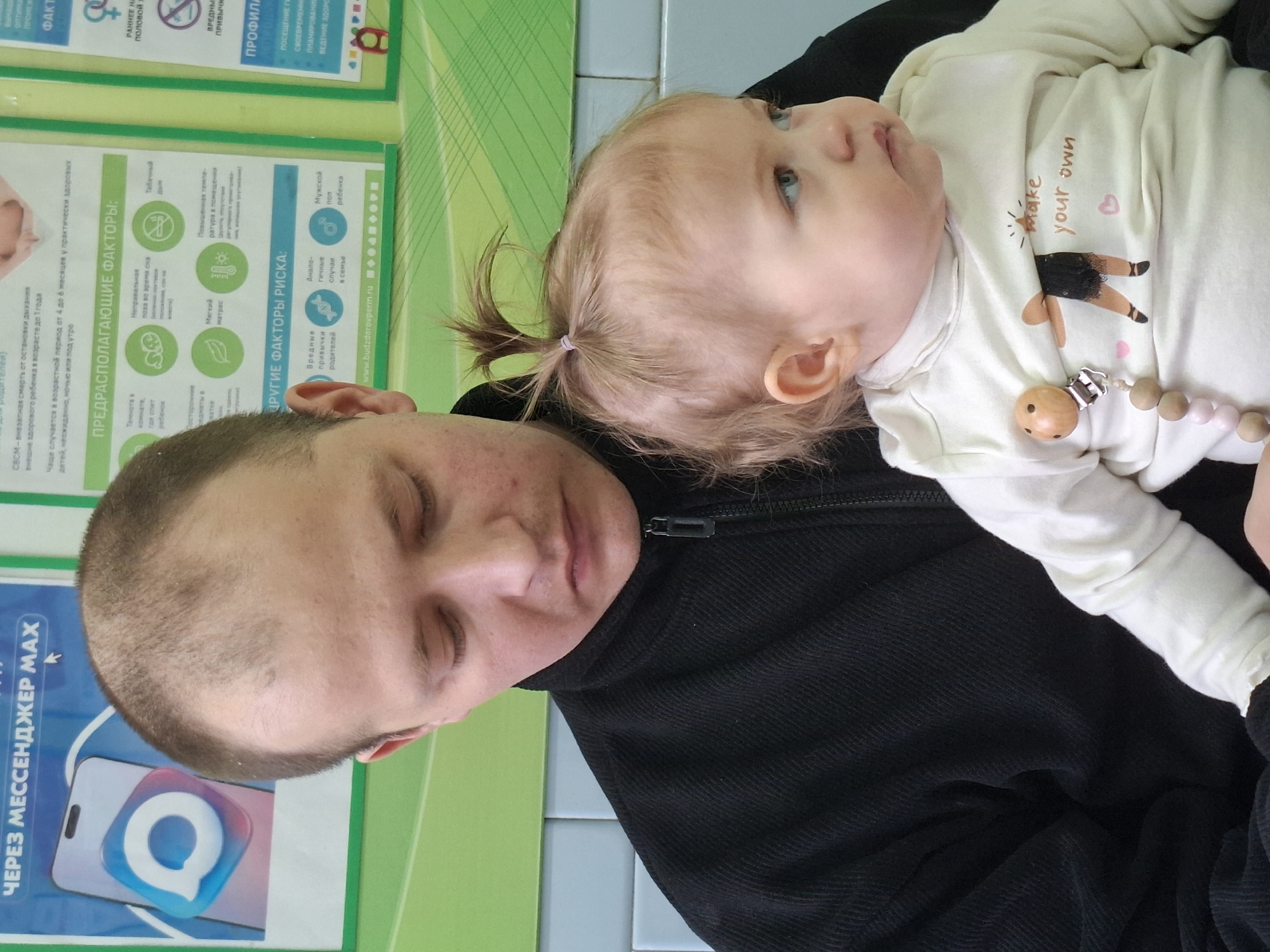A man is holding a baby in an indoor setting. The man is fair-skinned with short, thinning brown hair and closed eyes. He is wearing a black jacket. The baby has fair skin, light brown hair in a ponytail, and blue eyes. The baby is wearing a light-colored shirt with an illustration of a ballerina and the words "make your own" and a beaded pacifier clip. In the background is a green and blue sign in Russian; the lower left corner includes the words "ЧЕРЕЗ МЕССЕНДЖЕР MAX". The sign contains health and risk factor information. The location is most likely Chaikovsky, Russia.