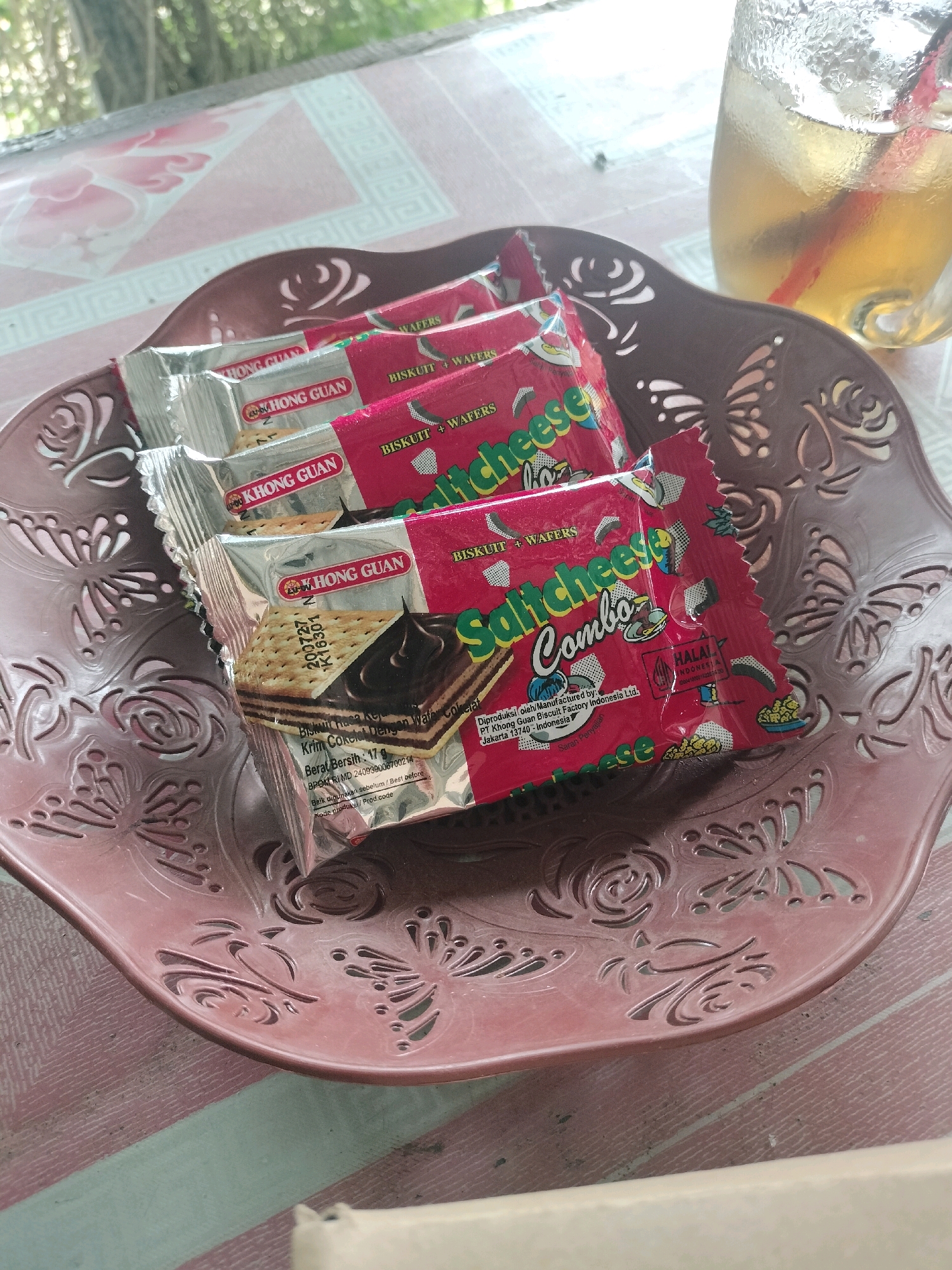 In Maburai, Indonesia, a brown, ornate plastic dish holds four individually wrapped packages of Khong Guan Saltcheese Combo biscuits. The biscuits are stacked on top of each other. The dish is placed on a table with a pink and white striped tablecloth. To the right of the dish, a glass of iced tea with a red straw is visible. The background shows a blurry outdoor scene with trees and foliage, suggesting a natural setting. The lighting is natural, indicating it is daytime.