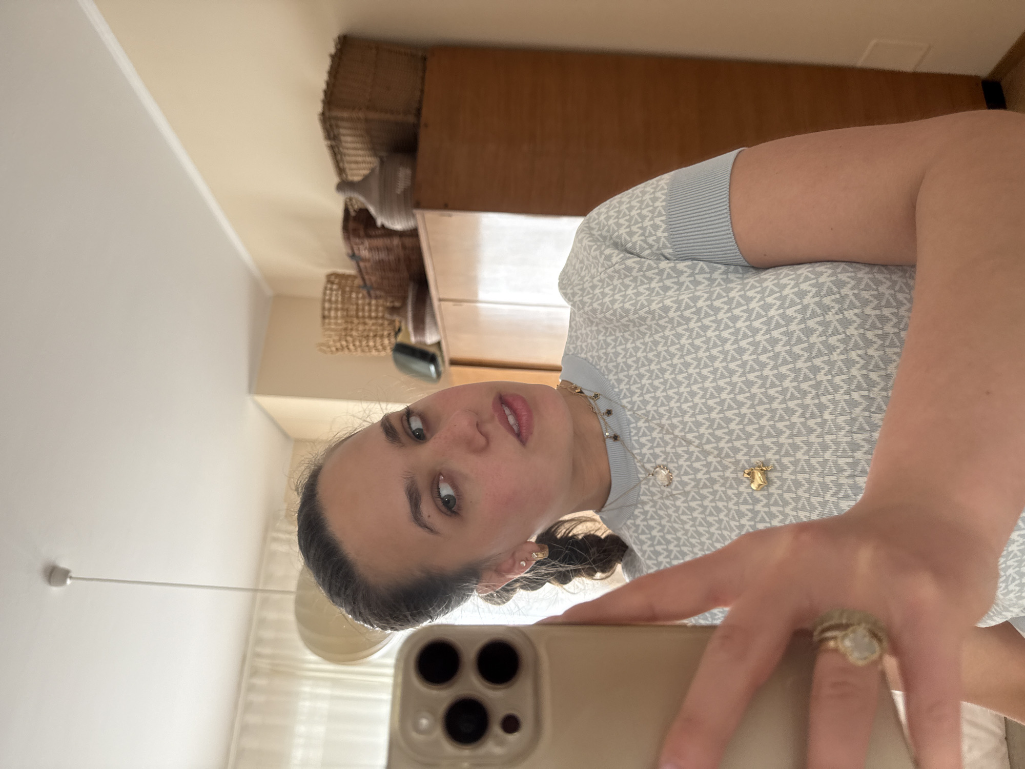 A woman is taking a selfie. The woman has brown hair styled in pigtail braids, fair skin, and is wearing multiple gold necklaces and rings. She is wearing a blue and white patterned shirt. The woman is holding a gold smartphone. Behind her is a cream-colored wall with a built-in wooden cabinet containing wicker baskets. The exact location in Milan, Italy cannot be determined from this image.