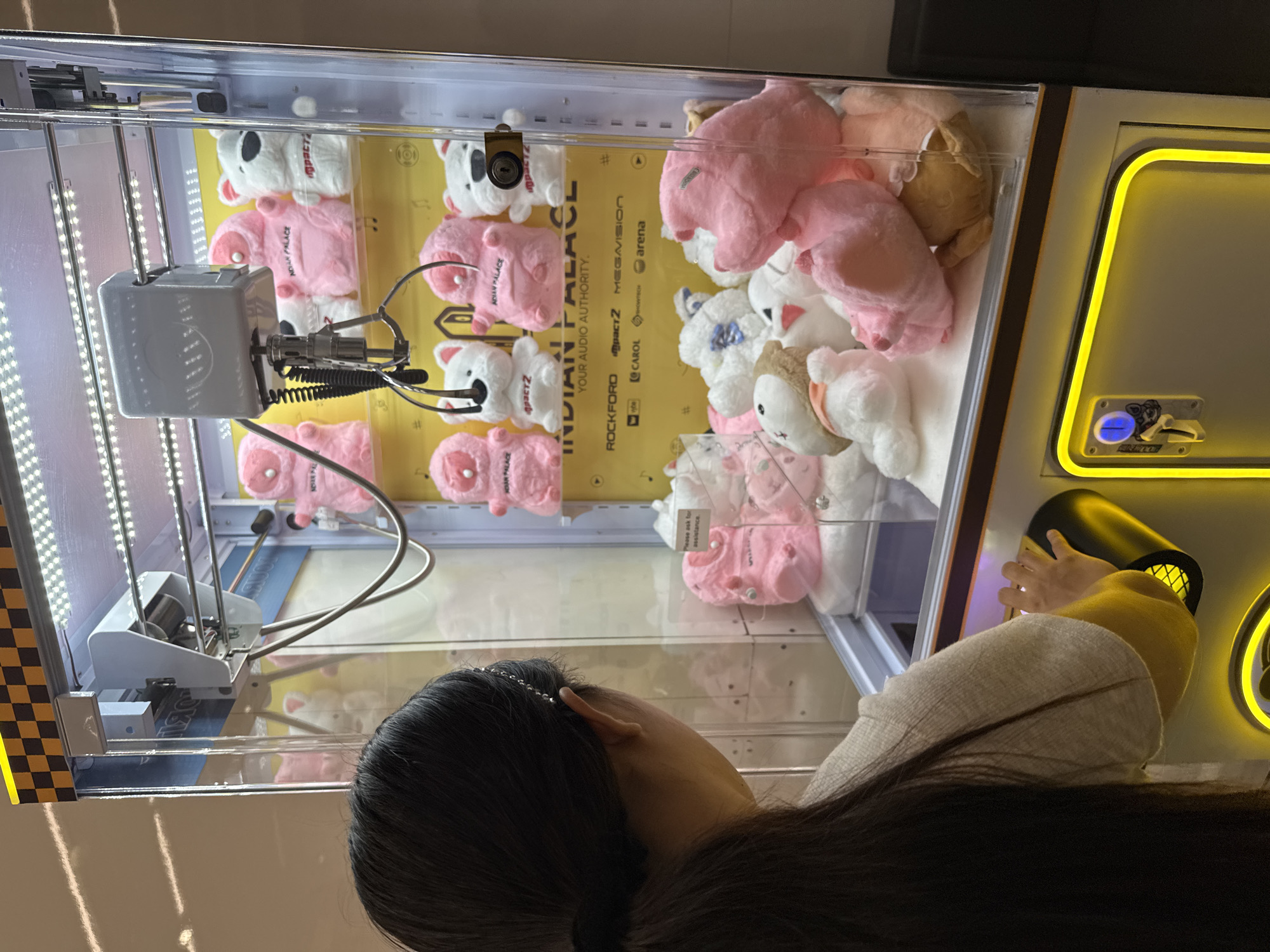 A person with long dark hair, visible from behind, intently gazes into a brightly lit claw machine. The machine features a transparent front and internal LED strip lighting, illuminating a dense collection of plush toys within. The majority of the stuffed animals are pink and white pig or bear-like figures, some adorned with the text "INDIAN PALACE" and "IMPACTZ" on their chests. A few brown and white animal plushies are also visible among them.
The claw mechanism, a silver-colored grabber, hangs suspended inside the machine, currently in an open position. On the right side of the machine, a vibrant yellow-lit control panel displays a blue button and a slot, likely for coins or prize dispensing. The person's hands are positioned near the machine, indicating engagement with the game, either playing or observing. A distinctive silver beaded hair accessory is visible in their dark hair.
The backdrop within the claw machine is a yellow panel, covered with various brand names and text, including "INDIAN PALACE," "YOUR AUDIO AUTHORITY.," "ROCKFORD," "MEGAVISION," "CAROL," and "arena." A small white tag inside the machine displays the message, "Please ask for assistance." The indoor setting suggests an arcade, entertainment center, or mall, likely in Davao City, Philippines. The scene captures a moment of focused interaction with an amusement game.