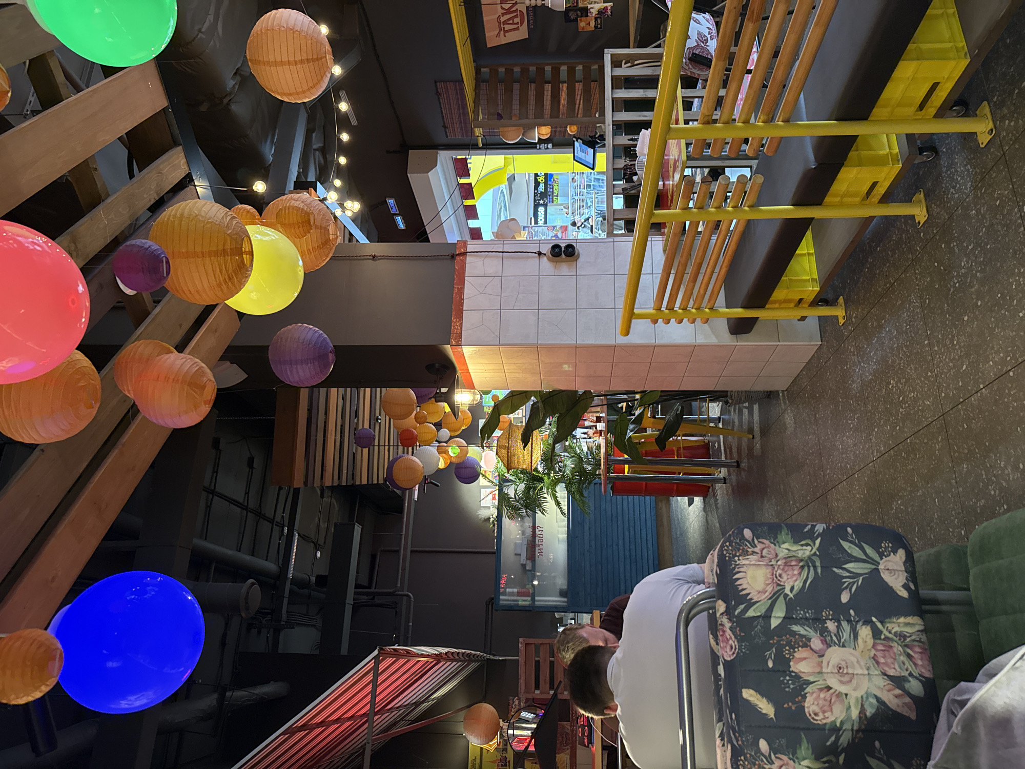 The image captures an indoor scene, likely a restaurant or cafe, with a vibrant, eclectic decor. Overhead, numerous colorful paper lanterns in shades of orange, yellow, pink, purple, and a striking blue illuminate the space, suspended from wooden beams. The walls are a mix of dark gray, white tiled sections with orange trim, and a section covered in patterned fabric. In the foreground, a man wearing a white t-shirt is seated at a table, his back to the camera. To his left, another person is partially visible. A dark fabric seat with a floral pattern is in the lower left corner. Further back, yellow benches with dark seating are arranged, supported by bright yellow posts and resting on yellow crates. A tropical plant is visible to the right, near a red metal barrel. In the background, a blue wall with shelving and a glass display case suggests a service or counter area. The flooring is a speckled gray tile. There is no indication of the city being Izhevsk, Russia, or any specific location.