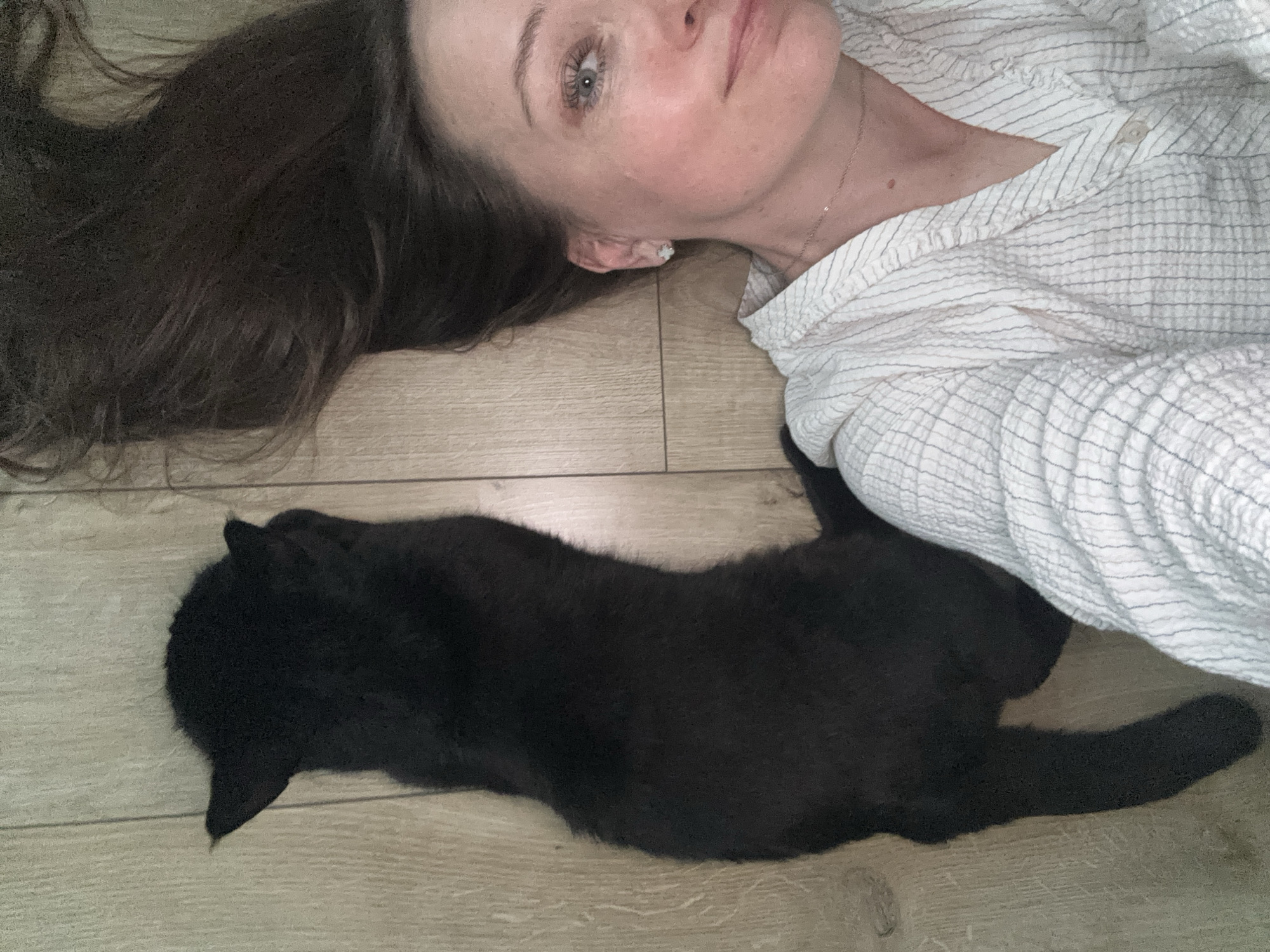 A woman lies on a light-colored wood floor next to a black cat. The woman has long dark hair, light skin, and is wearing a light-colored shirt with thin dark stripes. The cat is lying on its side, stretched out on the floor. The environment suggests an interior residential space, possibly in Chaikovsky, Russia.