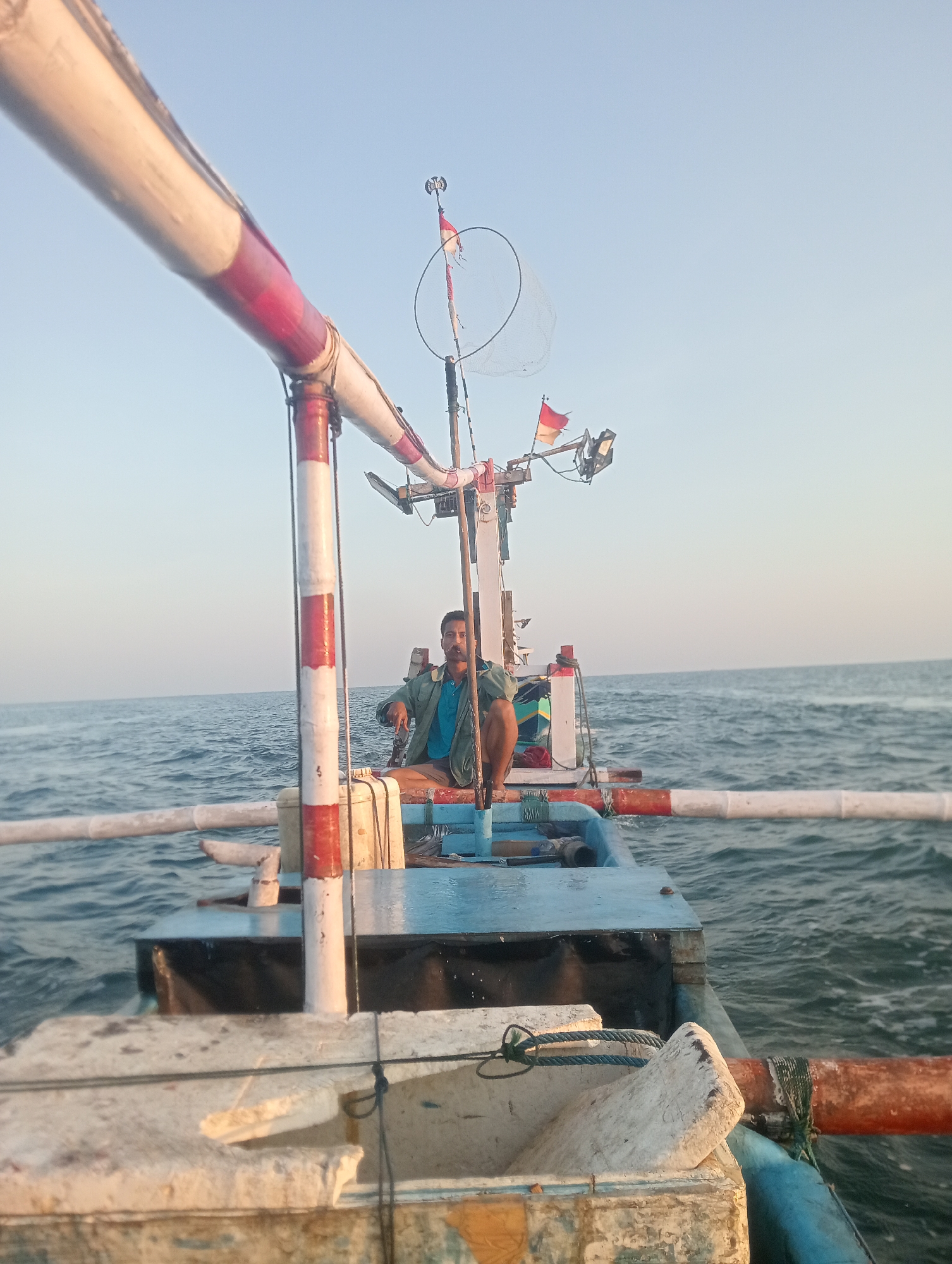 A man sits on a fishing boat at sea, likely near Pesisir, Indonesia, under a clear sky. The boat is equipped with a net and flags. The man, wearing a blue shirt and a green jacket, looks towards the camera. The water is calm, and the sun is low in the sky, casting a warm light. The boat is made of wood and has a white and red striped railing.