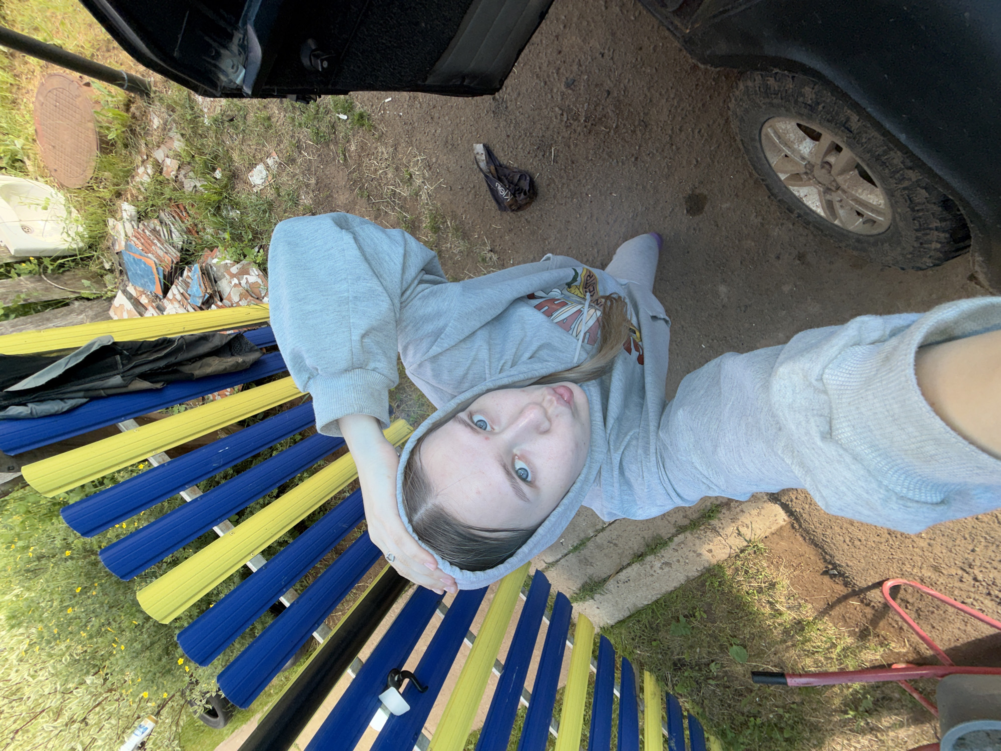 A young woman is featured in a high-angle selfie taken outdoors, likely in Zhigalki, Russia. She appears to be in her late teens or early twenties, with light-colored hair peeking from under a light grey hooded sweatshirt. Her blue eyes are directed towards the camera, and she has a playful expression with her tongue slightly extended. A ring is visible on her left hand.
The setting is an outdoor, somewhat disheveled area with a mix of dirt ground and patches of grass. To the left of the woman, a distinctive bench with alternating blue and yellow slats is visible. Beyond the bench, there is a metal manhole cover, a white basin-like object, and a pile of broken tiles, some of which are blue, scattered on the ground.
To the woman's right, a dark vehicle, possibly a truck or SUV, is partially visible, showing a dirty tire and part of its undercarriage. On the dirt ground near the vehicle, a small, dark, crumpled bag or item with what looks like "CK" or "CAL" text (possibly from "Calvin Klein") is discarded. Further right, a red metal frame or structure is partially seen. The overall lighting suggests it is daytime and sunny. The scene depicts a casual, candid moment in a semi-rural or rural environment.