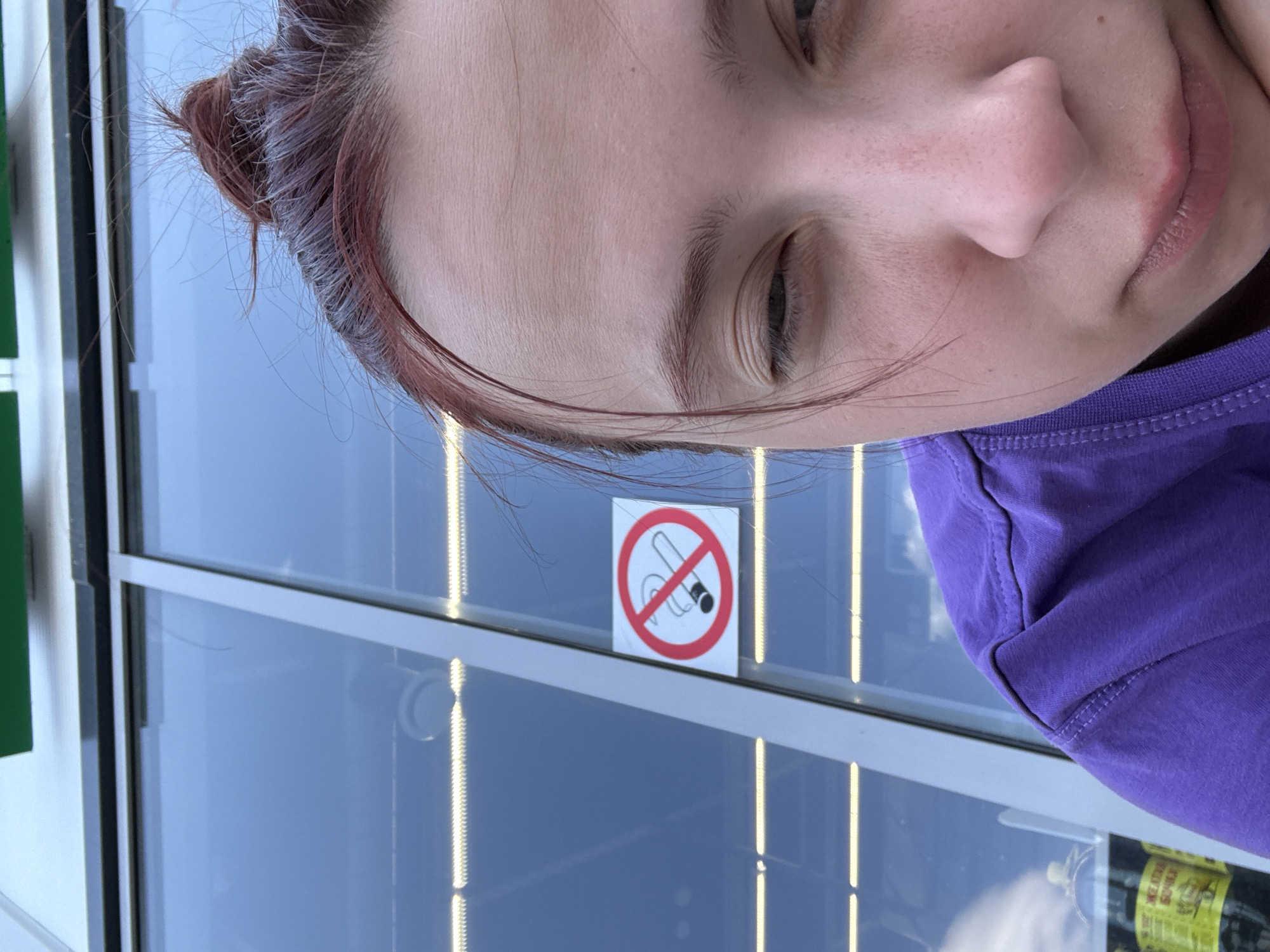 A close-up, slightly angled, indoor shot depicts a person with reddish-brown hair pulled back in a bun, wearing a purple shirt. Positioned behind the person and partially visible is a window with a "No Smoking" sign affixed to it. Based on the window's reflection, the exterior appears to be a bright, possibly overcast day. The specific location in Казань, Russia cannot be determined from this image.