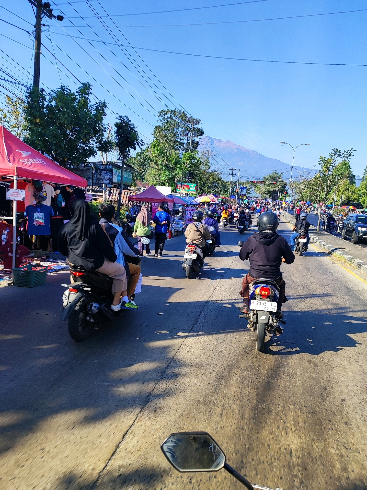 The image depicts a busy street scene, likely in Salatiga, Indonesia, on a clear, sunny day. The foreground is dominated by the perspective of someone riding a motorcycle, with a portion of their mirror visible at the bottom. Ahead, the road is filled with numerous motorcycles, many carrying two passengers, navigating through a bustling market area. Red and pink tents line the side of the road, displaying various goods, including clothing. In the background, lush green trees and distant mountains, possibly including Mount Merbabu, are visible under a bright blue sky. The scene suggests a vibrant local market or roadside fair, with a strong emphasis on motorcycle transportation, typical of urban Indonesian life.