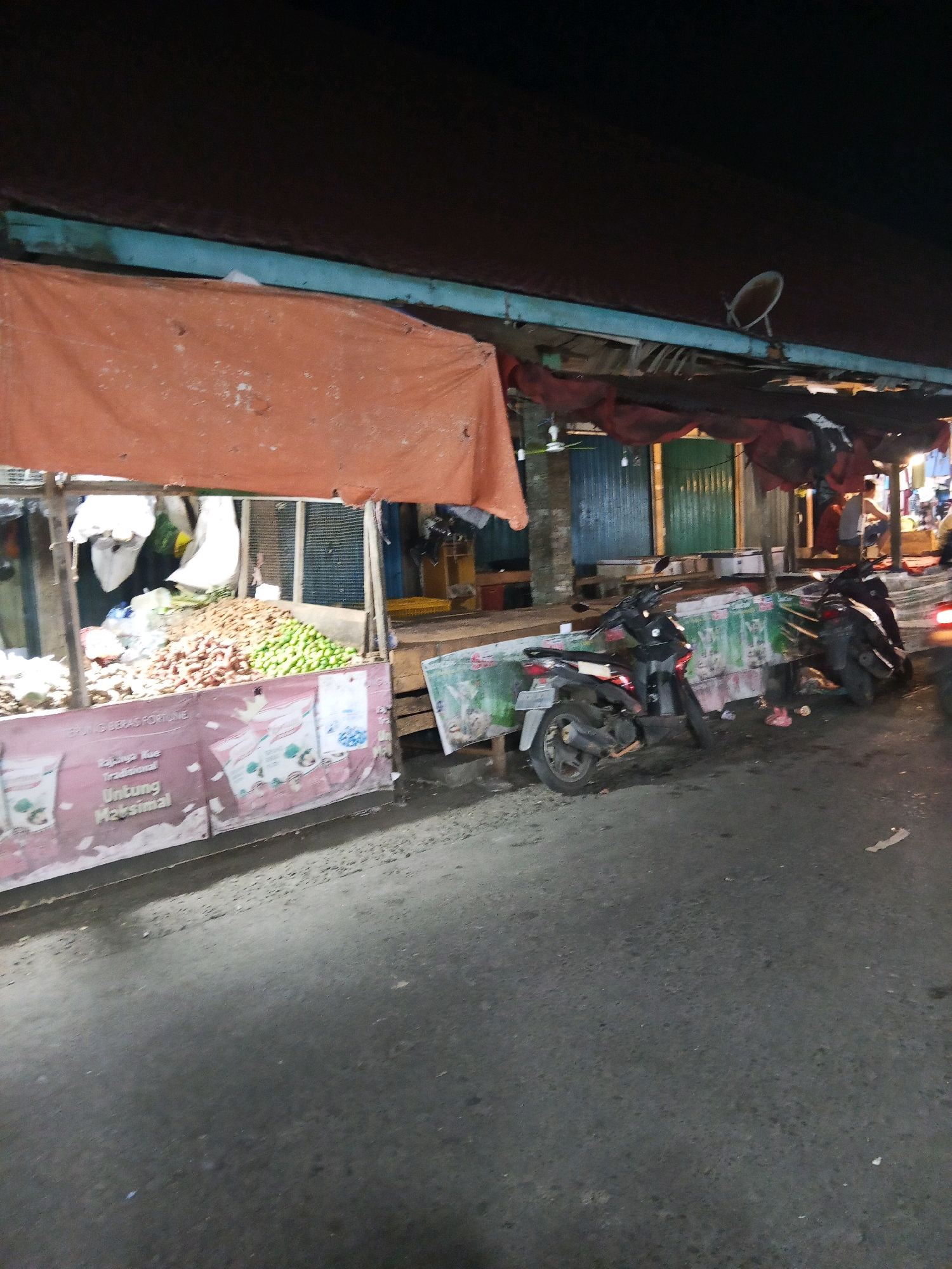 The image depicts a street scene at night in Lubuklinggau, Indonesia. In the foreground, a paved road surface is visible, with some scattered debris. To the left, a market stall is set up with a large orange tarp providing cover. Piles of produce, possibly fruits and vegetables, are displayed on the stall. The front of the stall features a large pink banner with text and images, including what appears to be product packaging.
Further to the right, two motorcycles are parked on the side of the road, partially in front of another market stall. This stall also has a covering, and further down the street, more market stalls and structures are visible. In the background, it appears to be nighttime, with artificial lighting illuminating the area. There are some indistinct figures of people in the background, suggesting an active market or street. A satellite dish is visible on a rooftop structure above the market stalls. The overall impression is of a local market operating after dark in a city setting.