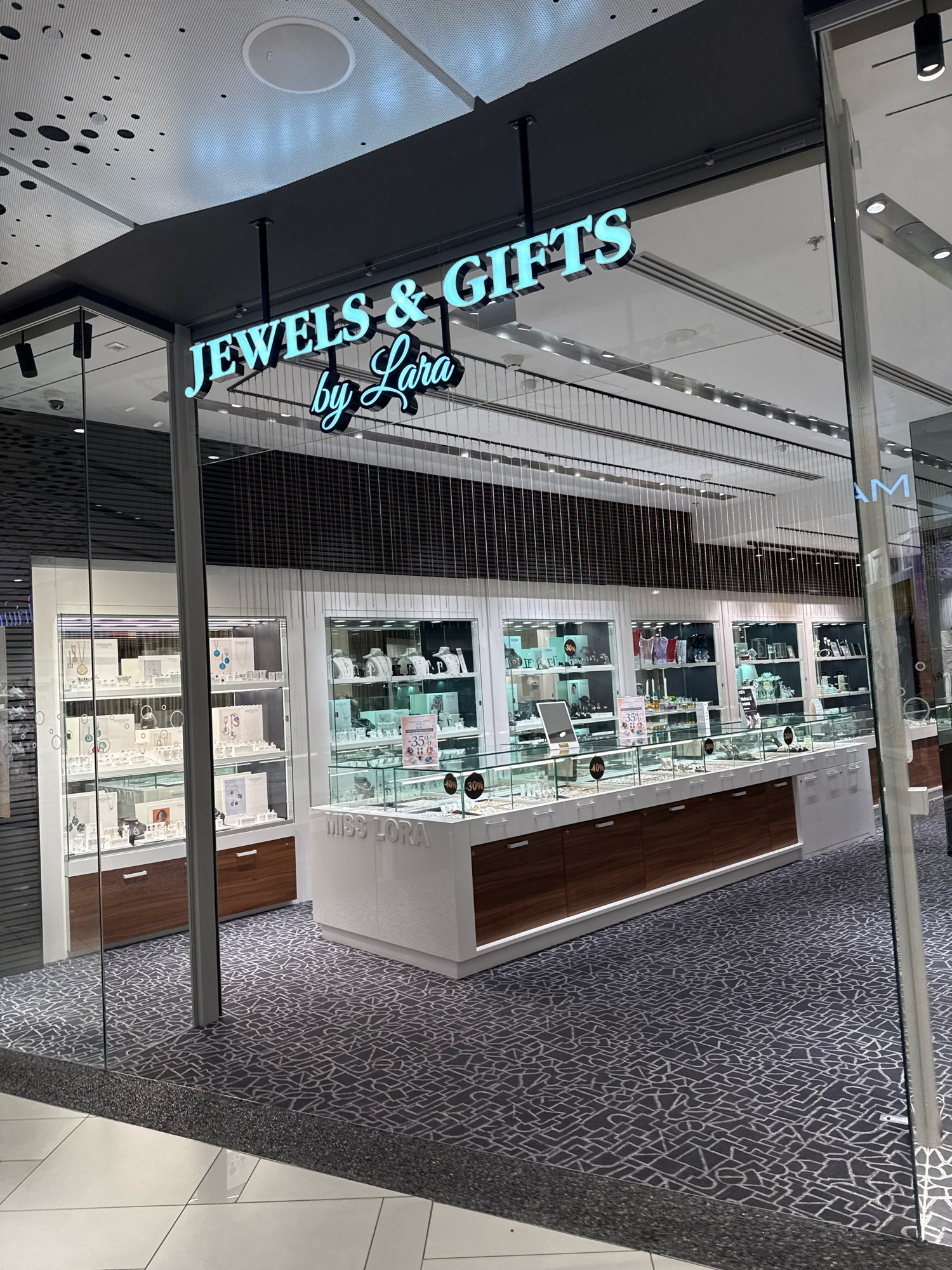 The image shows the exterior of a jewelry store called "JEWELS & GIFTS by Lara" located in Tallinn, Estonia. The store features a modern facade with a prominent illuminated sign. Inside, display cases filled with jewelry are visible. The store appears to be situated within a shopping mall, as indicated by the ceiling design and the presence of other stores in the background. The floor is covered with a patterned carpet. There are no people visible in the image.