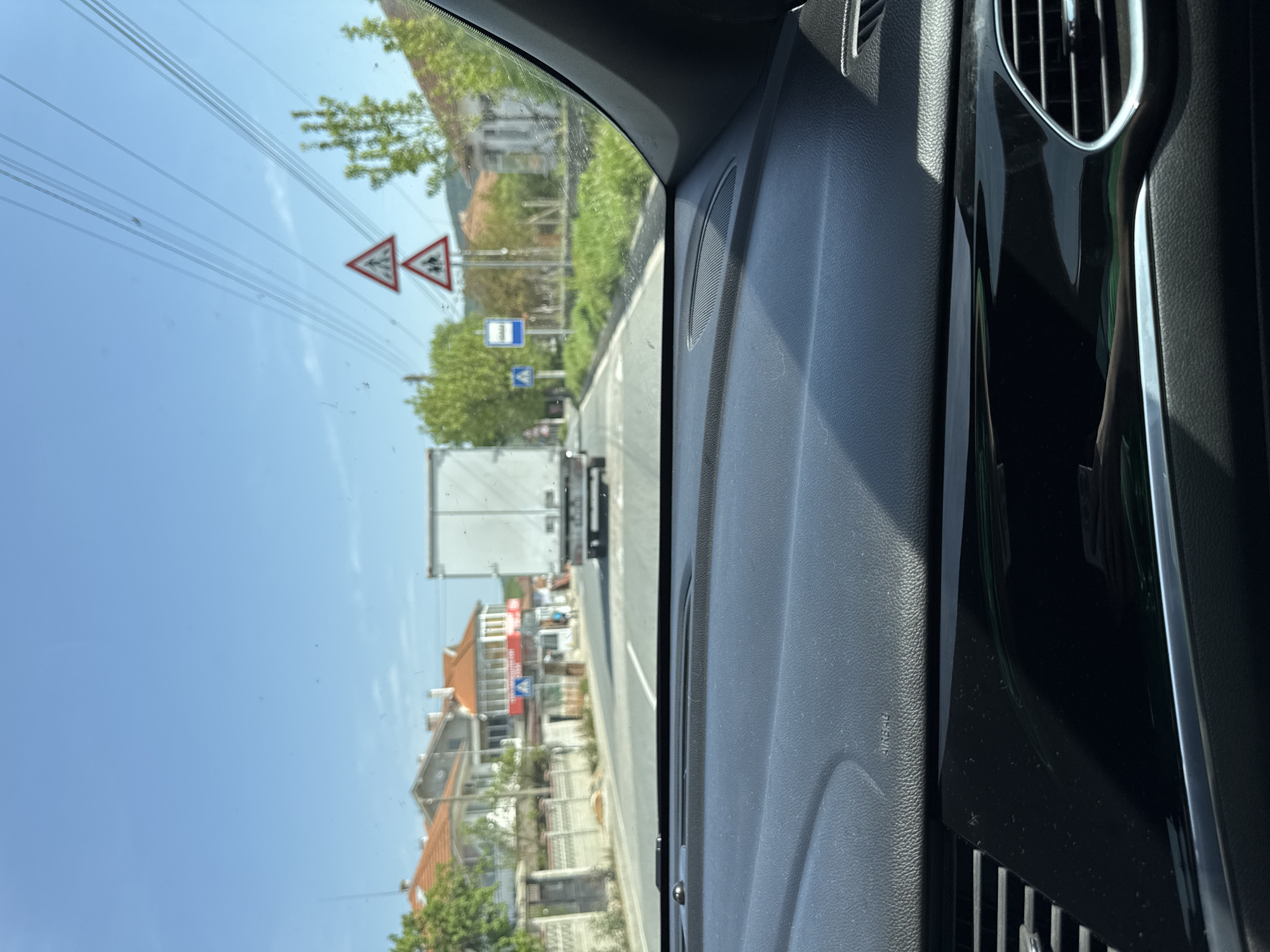 View from inside a vehicle, presumably a car, looking out the windshield. The vehicle is traveling on a road in what appears to be a residential or commercial area. A large white truck is visible in front, along with buildings on either side of the road. Road signs indicating pedestrian crossings and traffic conditions are present. The sky is clear and blue. The interior of the vehicle includes a dashboard with air vents and textured surfaces. The location is Daskotna, Bulgaria.