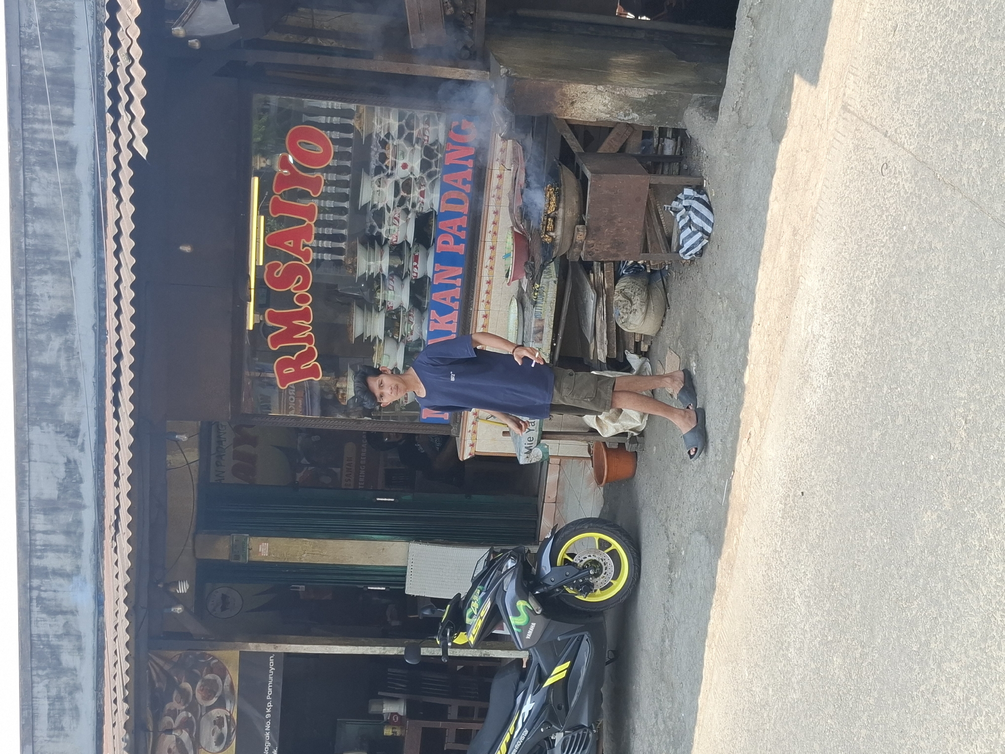 In Cibadak, Indonesia, a young man stands by his black Yamaha motorcycle parked on a concrete road in front of a restaurant. The restaurant has a large glass window displaying stacks of colorful bowls and a sign that reads "RM. SAIYO" in red lettering. Next to the restaurant, there is a cooking station with a wok over a fire, producing smoke. The man is wearing a dark blue t-shirt and khaki shorts, holding a cigarette in his right hand. The scene is bathed in sunlight, with shadows cast on the road. The environment appears to be a bustling street with a mix of commercial establishments.