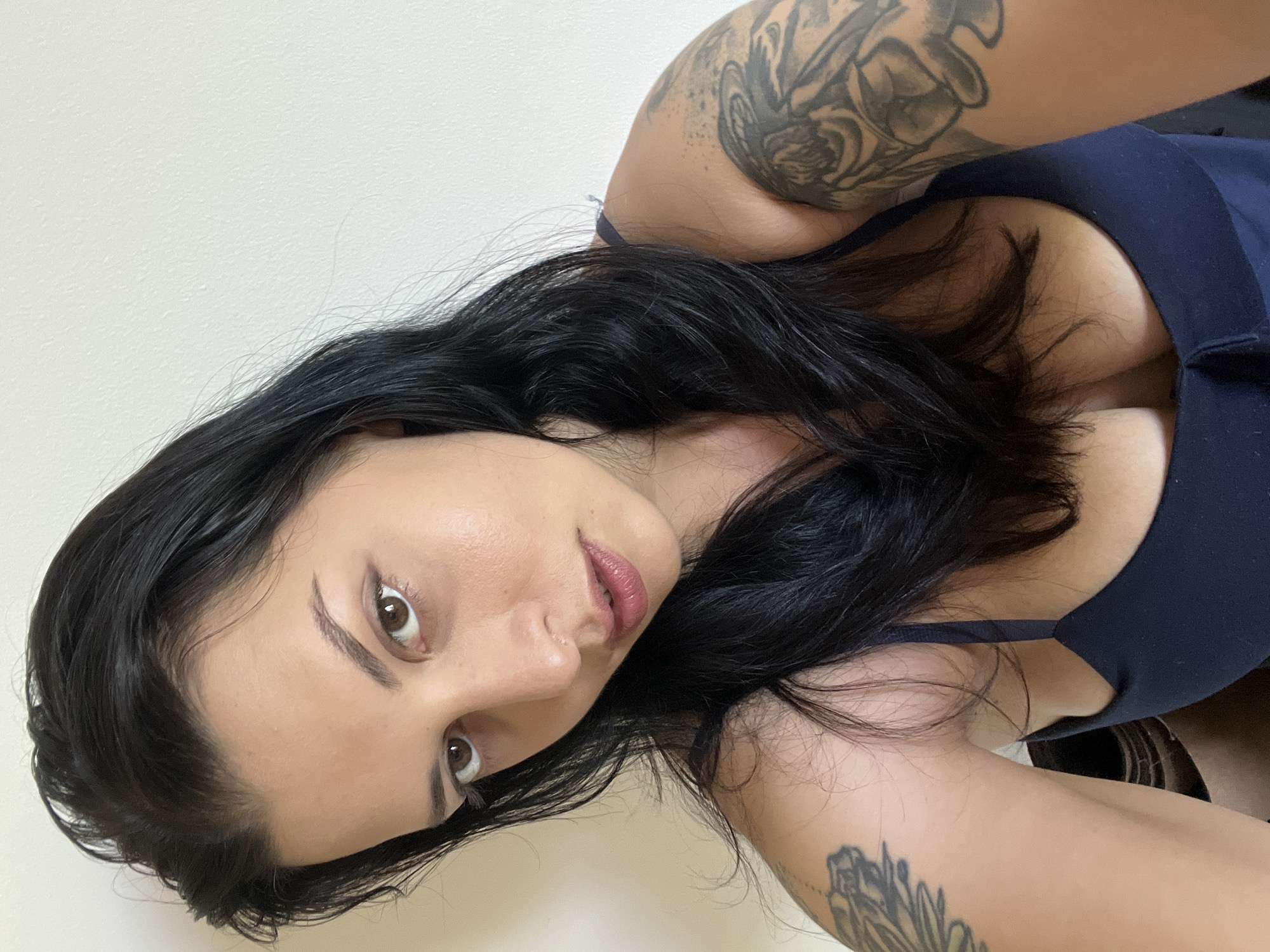 The image shows a close-up, selfie-style shot of a woman with dark hair and brown eyes. She is wearing a dark blue top, and has tattoos on her left arm and shoulder. The background is a plain, light-colored wall. There is no discernible location in the city of Đà Nẵng, Vietnam, nor any other specific environmental details. The image focuses entirely on the woman and her upper body.