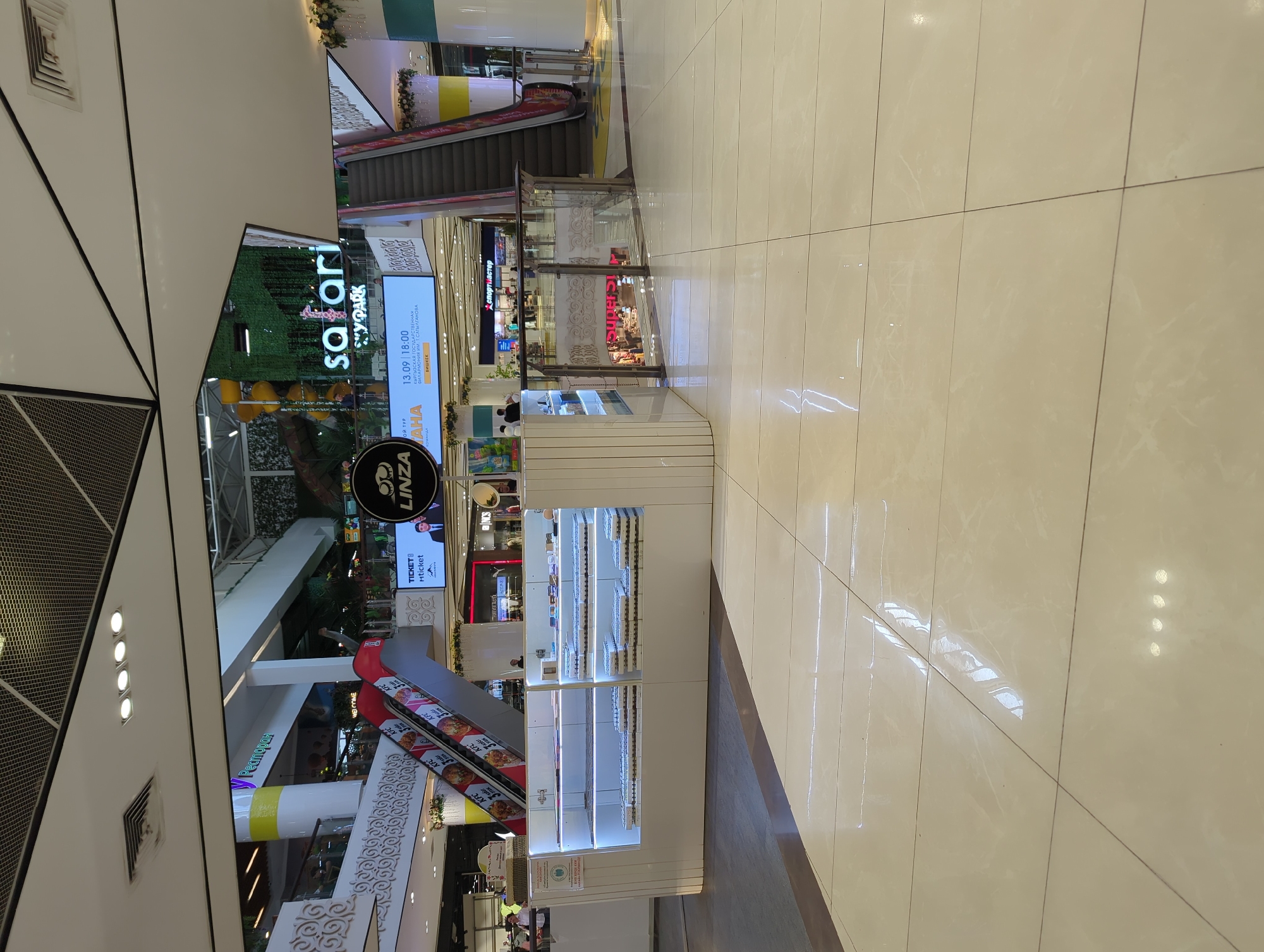 The image depicts the interior of a shopping mall in Bishkek City, Kyrgyzstan. The focal point is a display kiosk for the optician shop "LINZA," showcasing eyewear. Adjacent to the kiosk is an escalator and signage for "Safari YPark" and "KFC". Further back is a sign that says “Forever Baster” and “Supers”. The environment is brightly lit with polished tile flooring.