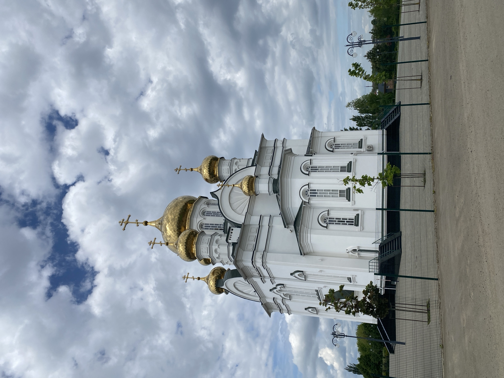 The image depicts the Cathedral of the Iveron Icon of the Mother of God, located in Bryukhovetskaya, Russia. The cathedral is white with gold onion domes, visible against a cloudy sky. A gray asphalt path runs along the right edge of the frame. A green fence separates the cathedral from the path, with small trees planted inside. Streetlights are visible beyond the fence line.
