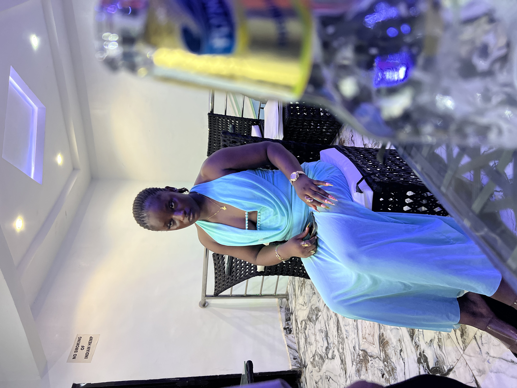 The image features a young woman seated indoors, possibly in a lounge or private area in Onitsha, Nigeria. The woman, with dark skin and braided hair, looks directly at the camera with a neutral expression. She is wearing a flowing, light blue or aqua sleeveless dress with a draped neckline. Her left wrist sports a gold watch, and her fingers are decorated with several gold rings and long, brightly manicured nails. She holds a dark-colored smartphone in her left hand.
She is seated on a dark, woven wicker-style armchair. In the blurred foreground, upper right, a reflective glass containing a drink is visible, suggesting a table nearby. The setting includes white walls, a mottled marble-patterned floor in shades of grey and white, and a modern ceiling with recessed lighting and an illuminated, rectangular cut-out emitting a purple glow. The artificial lighting suggests an evening or nighttime scene.
A white sign with black text is mounted on the wall to the left of the woman, clearly stating: "NO SMOKING OF INDIAN HEMP". The overall ambiance indicates a contemporary indoor venue.