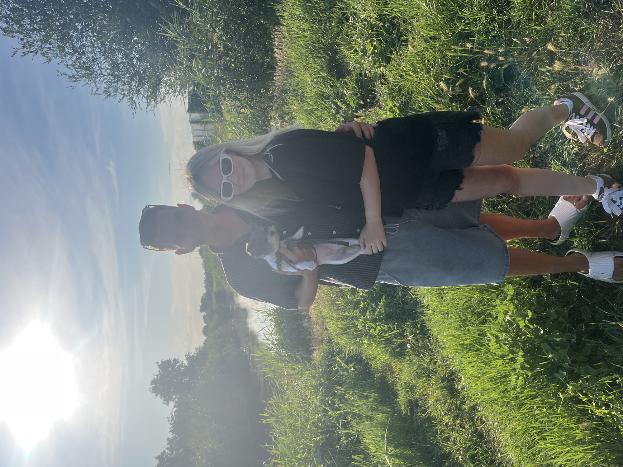 A young couple, a male and a female, are standing in a grassy field near a body of water, possibly a canal or river. The male, on the left, is wearing a striped short-sleeved shirt, grey shorts, and white slides. He is holding a small dog in his arms. The female, on the right, is wearing a black t-shirt and black denim shorts. She has blonde hair and is wearing white sunglasses. The sun is shining brightly, casting a warm glow over the scene. The background consists of lush green grass, trees, and the water. The overall impression is a casual, outdoor moment. The location is identified as Yanovo-Grushevskiy, Russia.