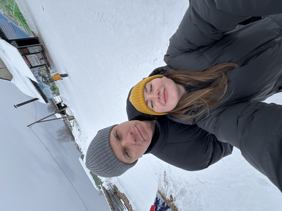A selfie image depicts two individuals in a snowy outdoor environment identified as the Zaosinovo Ostrich Farm in Zaosinovo, Vologda Oblast, Russia. In the foreground, a male subject wears a grey knit beanie and a black puffer jacket, looking directly at the camera with a neutral expression. Positioned in front of him and to the right, a female subject wears a yellow knit beanie and a black puffer jacket; her eyes are closed, and she displays a slight smile. The ground is uniformly covered in deep snow under an overcast grey sky. In the mid-ground, a wooden utility pole with overhead lines is visible. To the right, a building with a snow-laden roof features multiple signs in Russian, prominently displaying "СТРАУСИНАЯ ФЕРМА" (Ostrich Farm), "ЭКСКУРСИИ" (Excursions), and a phone number (278 15 36). A small individual in a bright yellow jacket is walking away from the camera, further down a snowy slope toward this building. A partially obscured blue and white patterned object rests on the snow near some dry vegetation on the far left.