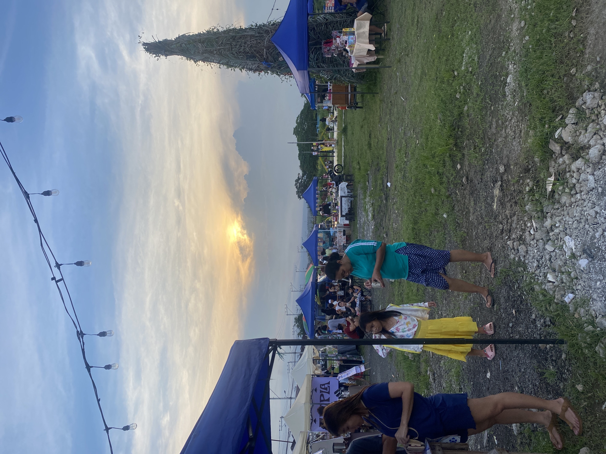 This image captures an outdoor market or festival in Davao City, Philippines, during the late afternoon or early evening. The sky displays a beautiful transition from a clear blue on the left to warm, golden and orange hues on the right, indicating either a sunset or sunrise, with scattered clouds adding texture.
In the foreground, a string of bare light bulbs hangs across the upper left, suggesting preparations for evening activities. Several people are present, moving casually through the scene. A woman in a blue top and dark shorts is seen from behind in the lower foreground. To her right, a young girl in a yellow skirt and a white patterned top looks down. Further back, a man in a green t-shirt and patterned shorts stands, holding an object in his hand.
The event takes place on an open ground with patches of grass, dirt, and some rocky areas. Numerous temporary structures, including blue and white canopy tents, are set up in rows, likely hosting vendor stalls or activity areas. Under one of the white tents in the mid-ground, the word "UTOPIA" is partially visible. People are seen gathered around these stalls, browsing, or conversing.
A very notable feature is a tall, conical structure on the left side of the mid-ground, crafted from organic materials like woven branches or leaves, resembling a large, rustic monument or a unique Christmas tree. The overall ambiance is relaxed and community-oriented, with the warm natural light enhancing the festive feel.