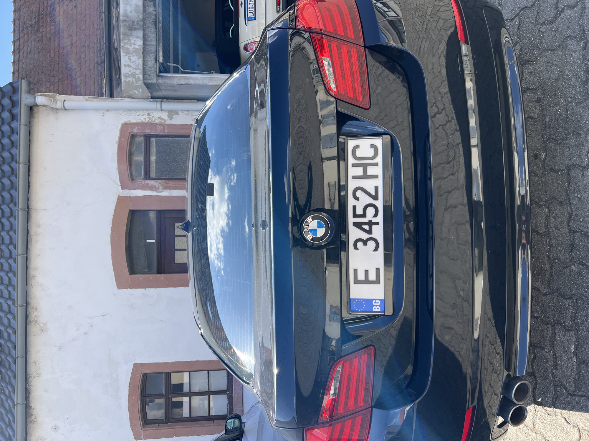 The image shows the rear of a dark-colored BMW, specifically its license plate with the number "E 3452 HC". The license plate also has the "BG" country code, indicating that the car is registered in Bulgaria. The car is parked on a paved surface, possibly a street or parking lot. In the background, there is a white building with several windows. The building appears to be an older construction with some visible wear and tear. The sky with clouds is reflected in the car's rear windshield. The location is not identifiable based on the image content.