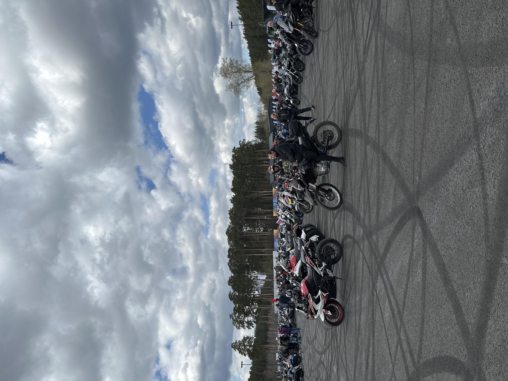 The image shows a gathering of motorcycles in a large paved area, likely in the city of Yekaterinburg, Russia. Several rows of motorcycles are parked, with people standing around some of them. Tire tracks cover the asphalt surface. A line of trees is in the background and a cloudy sky is overhead.
