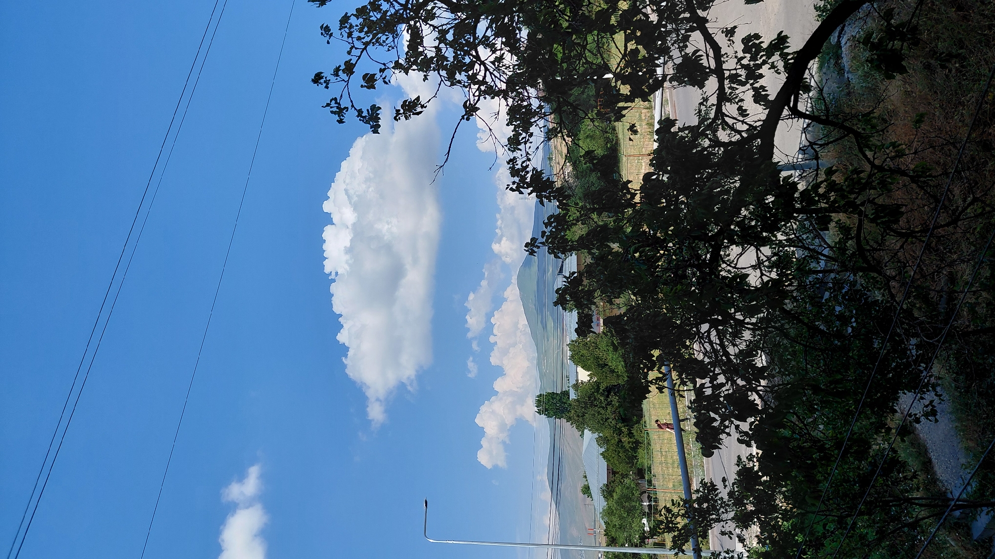 The image captures a bright, sunny day with a clear blue sky and scattered white clouds. In the foreground, dark, leafy branches of a tree obscure part of the view, suggesting a perspective from a window or balcony. Below the tree canopy, a street is visible, with a light pole standing to the left. Further in the distance, there are buildings with light-colored roofs, surrounded by lush green trees. A gentle, rolling mountain range forms the backdrop, its slopes covered in vegetation under the expansive sky. In the mid-ground, a person is walking along a path adjacent to the road. The overall impression is one of a peaceful, rural or semi-rural setting, likely in or around Abovyan, Armenia, given the characteristic landscape and architecture.