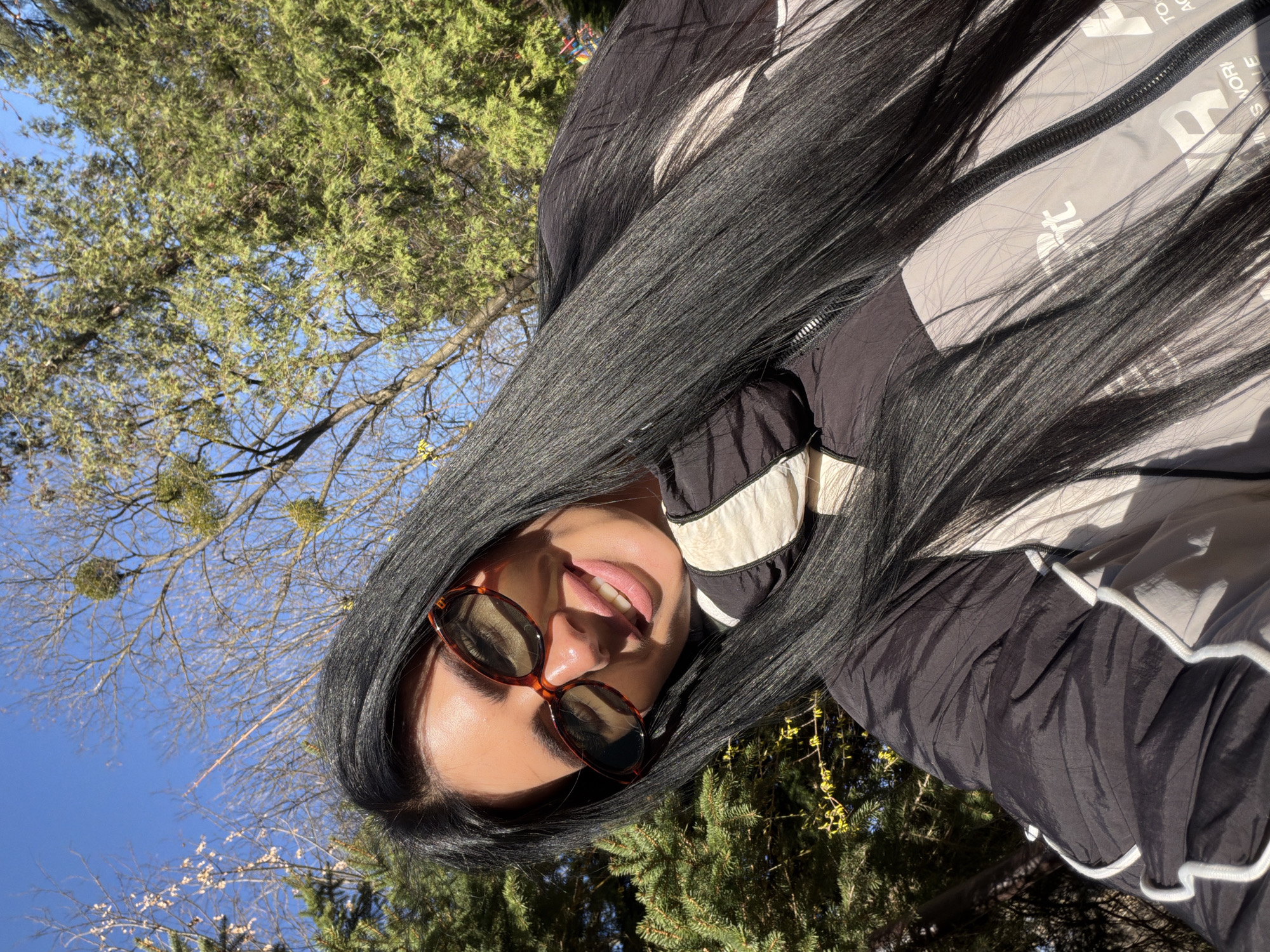 A woman with long, dark hair and wearing sunglasses is outdoors. She is wearing a black and grey jacket. The background includes green trees, branches and a clear blue sky. Due to the lack of contextual information, a specific location in Hrodna, Belarus cannot be determined.