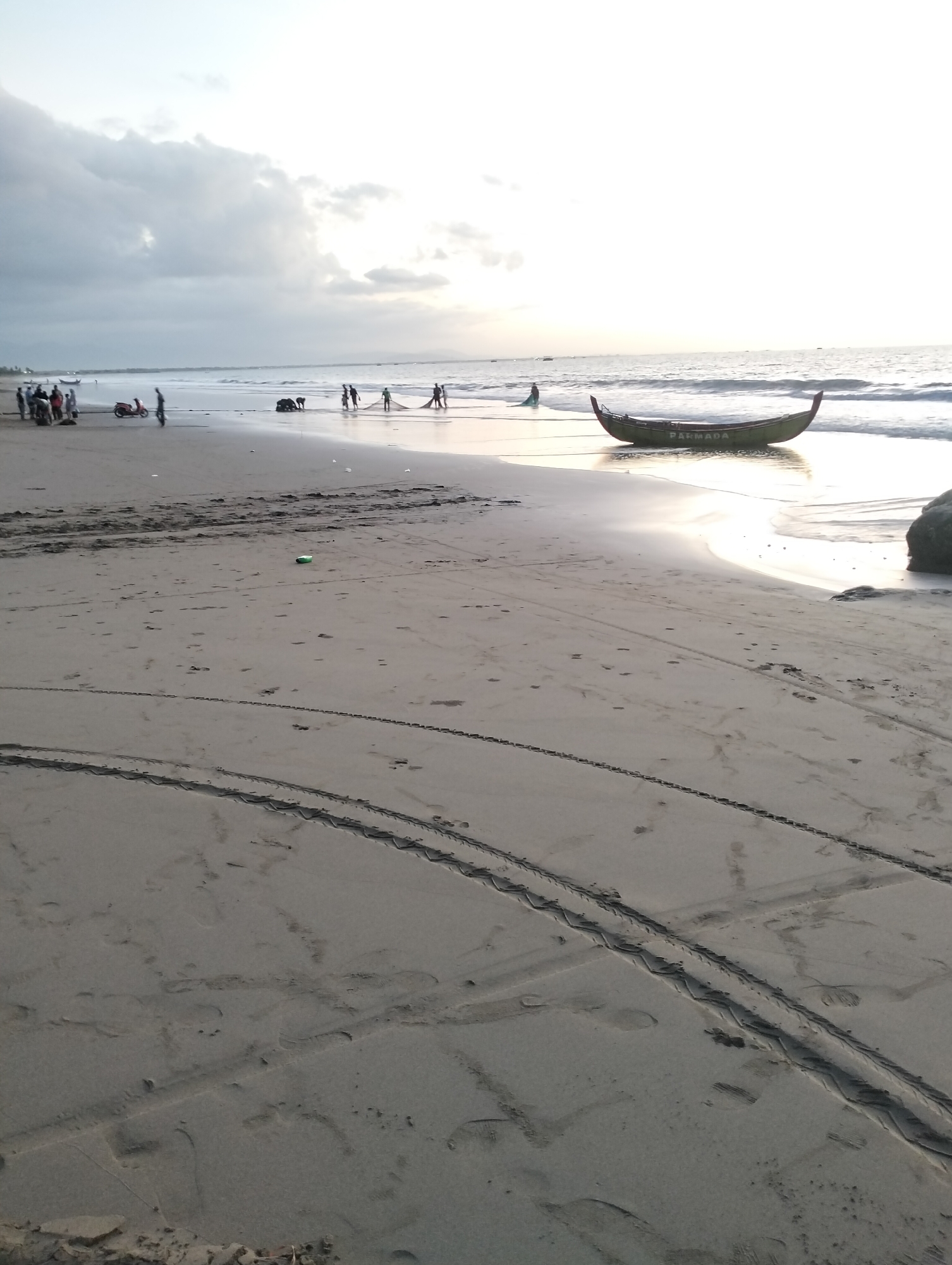 The image depicts a beach scene in Peuneulet Baroh, Indonesia, under a cloudy sky. In the foreground, the sandy beach is marked with tire tracks and footprints. A small green object, possibly a bottle, rests on the sand. Further down the beach, near the water's edge, a group of people are engaged in activities. Some are gathered around what appears to be a fishing net, while others stand by a motorcycle. A traditional fishing boat, labeled "PARMADA," is partially submerged in the shallow water. The ocean stretches out to the horizon, with gentle waves lapping the shore. The overall impression is one of a working fishing community at dusk.