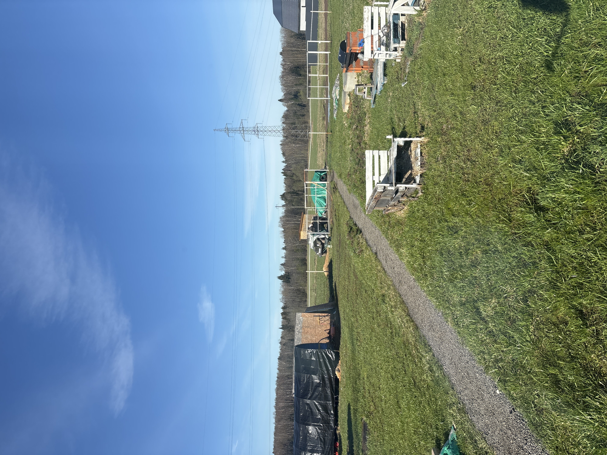 The image shows a landscape in Раздолье, Russia. Overhead power lines run across the scene, supported by a metal tower. The ground is covered in green grass, with a gravel path cutting through the field. Various objects are scattered along the path, including wooden structures, a covered trailer, and construction materials. The sky is blue with scattered clouds.