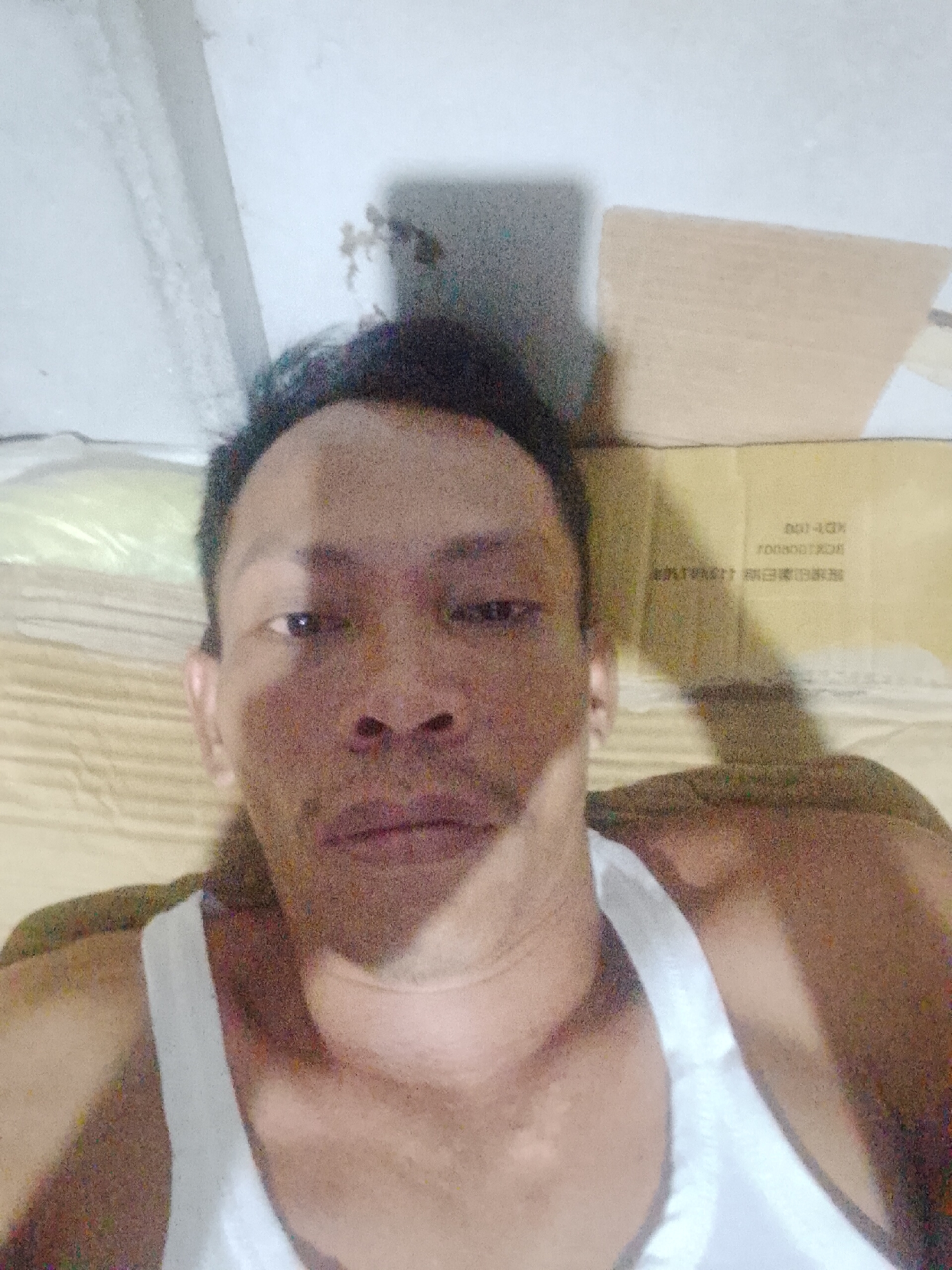 A man is lying down in a location identified as Bukit Ketok, Indonesia. He is wearing a white tank top. Behind him is a cardboard box with printed text in what appears to be a non-Latin script, possibly Japanese or Chinese. The immediate environment appears to be a room with a light-colored wall.
