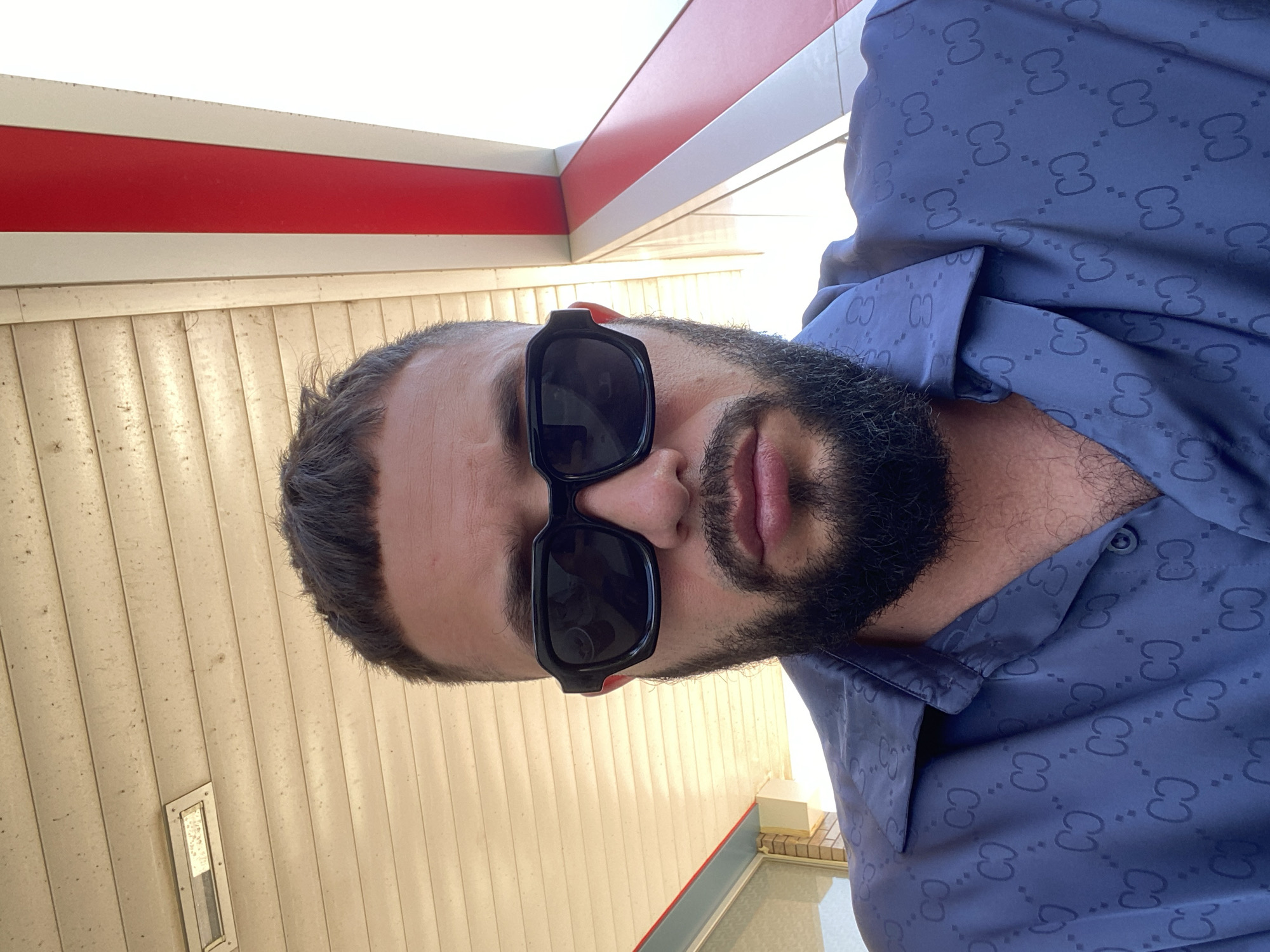 A man with short dark hair, a beard, and sunglasses is pictured from the chest up. He is wearing a blue button-down shirt with a repeating dark pattern. The background features a beige, corrugated ceiling with a light fixture. To the right, a red and white striped structure is visible, suggesting an outdoor setting. The image appears to have been taken at a gas station in Korkmaskala, Russia.