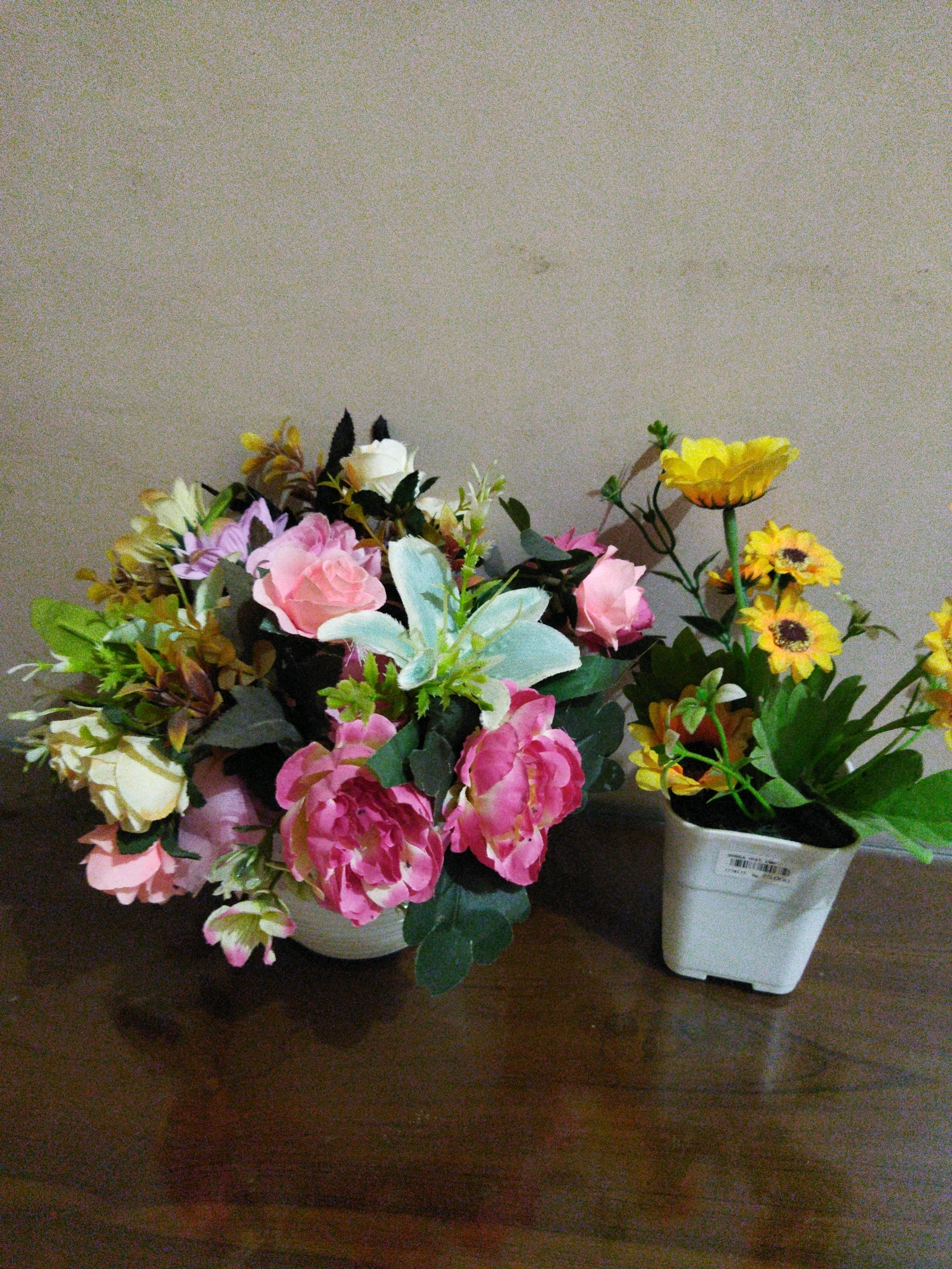 The image displays two floral arrangements on a wooden surface against a textured, light-colored wall. The arrangement on the left is larger and appears to be in a white, rounded vase, filled with a mix of pink, white, and yellow flowers, along with greenery. The arrangement on the right is smaller, in a white square pot, and features yellow flowers resembling sunflowers and gerbera daisies, along with green leaves. A shadow of the yellow-flowered arrangement is visible on the wall behind it. The scene appears to be an indoor setting, possibly a home or a shop, located in Banjar Agung, Indonesia. There are no people or animals visible in the image.