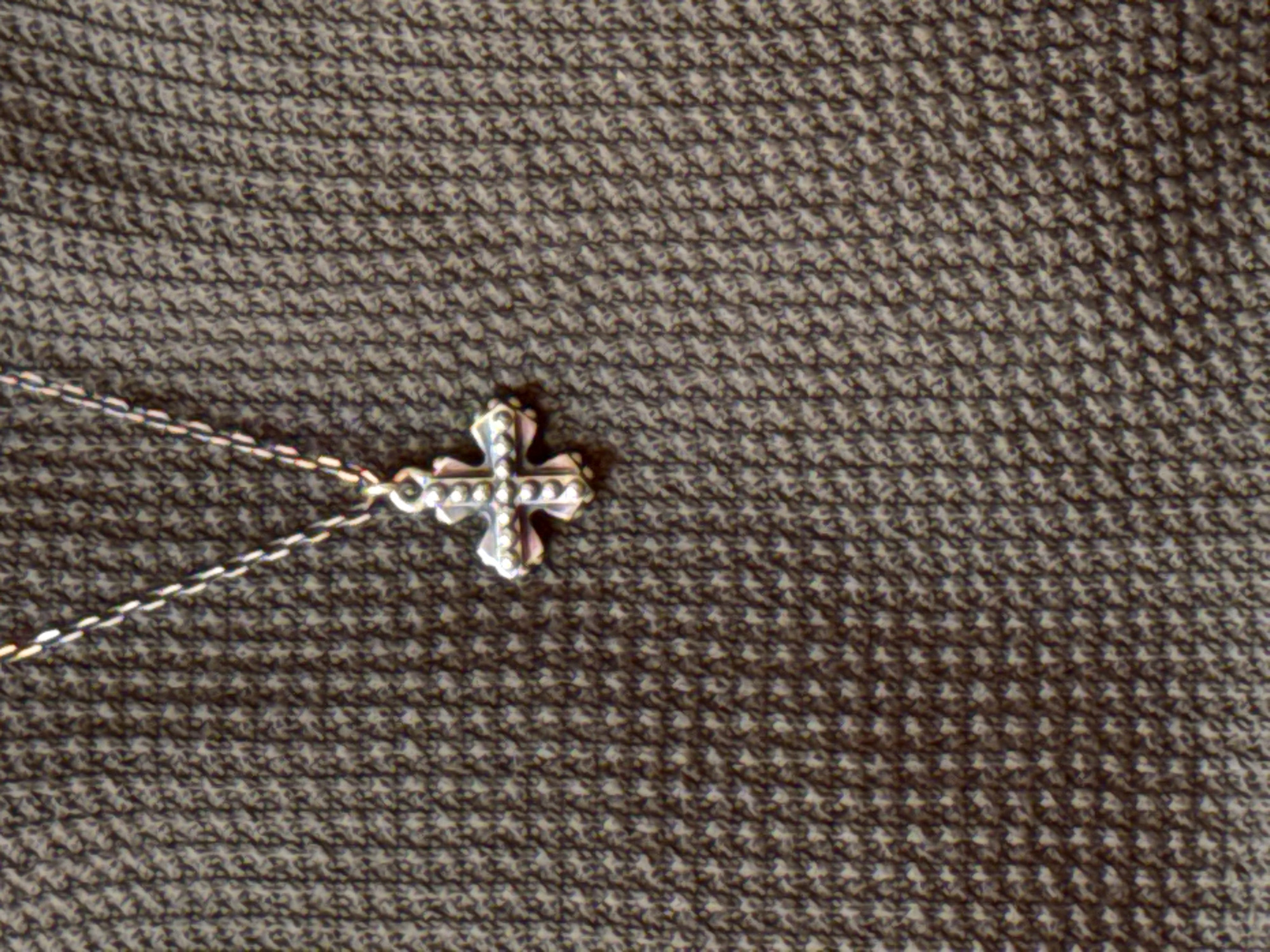 The image shows a close-up of a silver cross necklace resting on a textured brown fabric. The cross is ornate with intricate details and appears to be a pendant on a delicate silver chain. The fabric has a woven pattern, possibly houndstooth or a similar small, repeating geometric design. The lighting suggests an indoor setting. There is no indication of the city of Yerevan, Armenia, in the image.