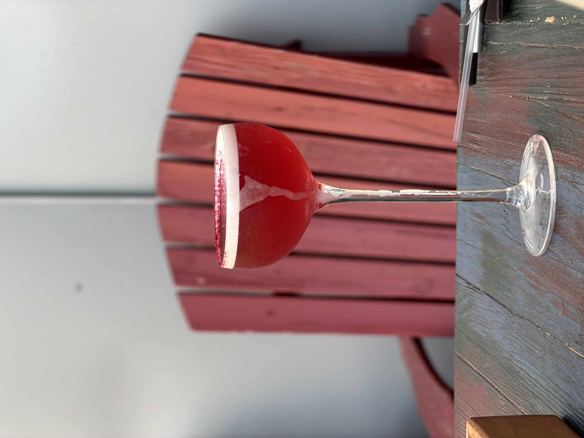 A close-up, eye-level shot showcases a vibrant red cocktail in a coupe glass. The drink is topped with a frothy white foam and garnished with a dusting of red powder around the rim of the glass. The cocktail is positioned on a weathered, multi-colored wooden table. In the background, a red wooden Adirondack chair with vertical slats is visible, suggesting an outdoor setting. The overall lighting indicates daytime, with diffused sunlight. The location is identified as Izhevsk, Russia.