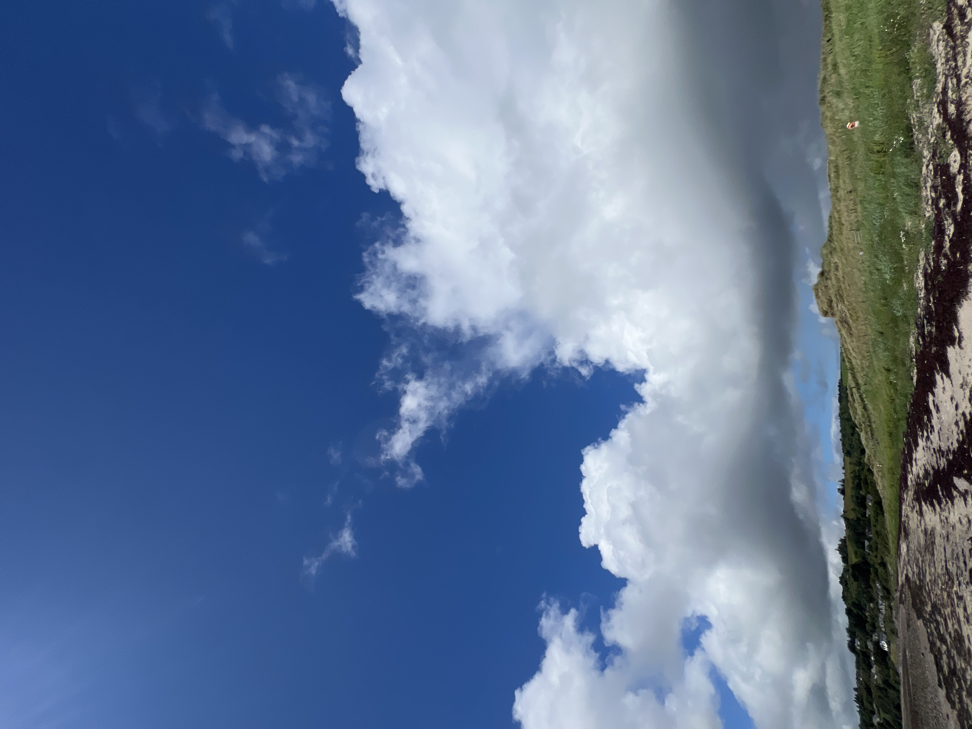 This media file depicts a dramatic sky over a coastal landscape, likely Mountcharles, Ireland. The scene is dominated by a vast expanse of deep blue sky, interrupted by a striking contrast between bright, fluffy white clouds and dark, ominous storm clouds. The clouds appear to be moving, creating a sense of dynamic change in the atmosphere.
The lower portion of the image shows a sloped grassy area with some vegetation, suggesting a hillside or dune. A rough, dark path or surface, possibly sand or wet earth with dark debris, runs along the right edge of the frame. In the distance, a line of trees and what appear to be small buildings or houses can be seen along the base of the hill, indicating a settled area within the natural landscape. A small, bright orange object, possibly a buoy or life preserver, is visible on the grassy slope, a solitary detail against the backdrop of the weather.
The time of day is not precisely clear, but the quality of light suggests it could be late afternoon or early evening, given the potential for dramatic cloud formations. The weather is clearly mixed, with sunshine implied by the bright white clouds, but the presence of the dark storm clouds suggests the possibility of rain or a change in conditions. The overall mood conveyed is one of natural beauty and the power of the elements, with a hint of the pastoral from the distant settlement. There is no visible text in the image.