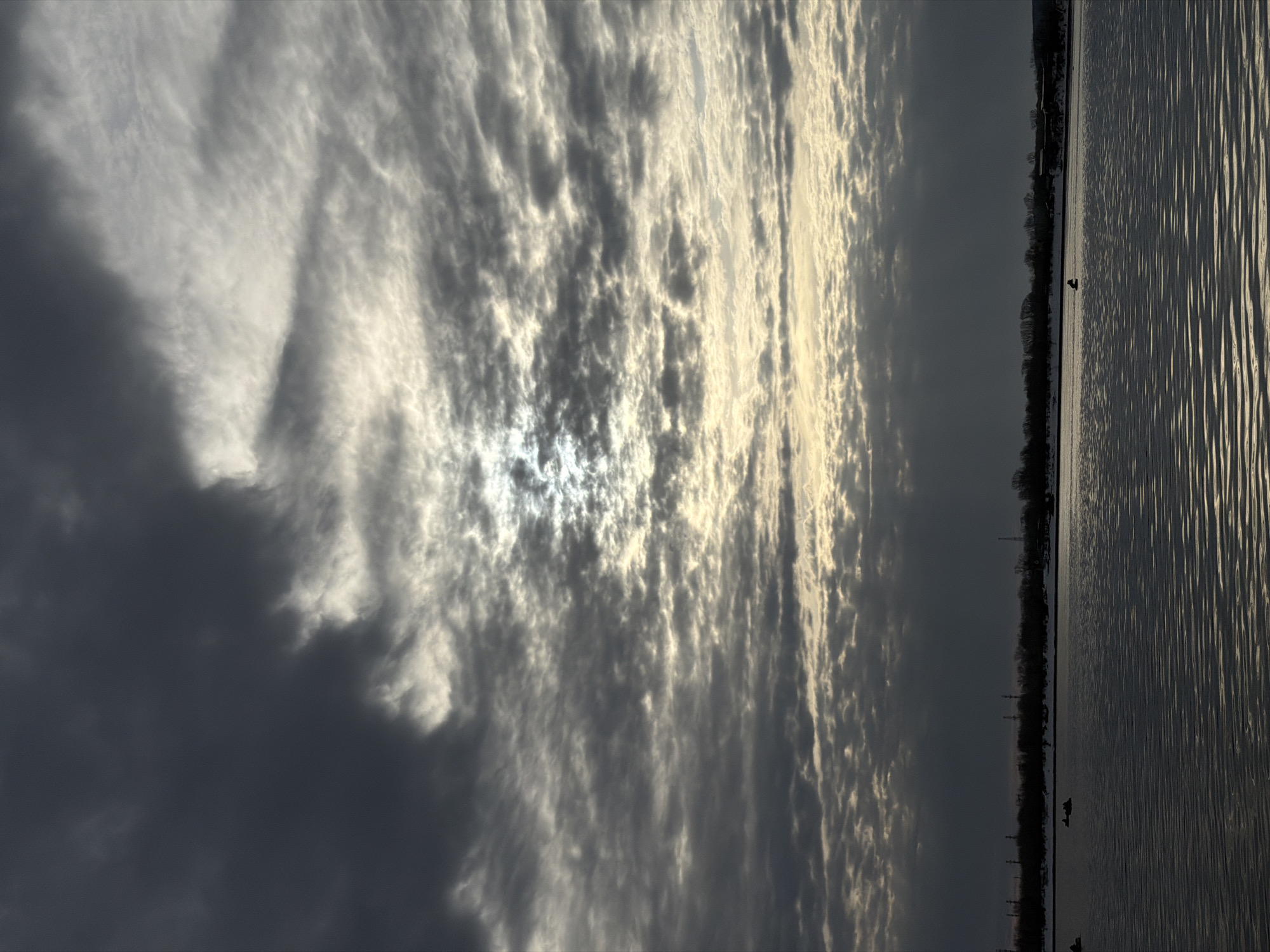 The image depicts a body of water adjacent to a forested shoreline beneath a sky filled with clouds. The water surface shows a rippled texture, reflecting light in a striped pattern. The shoreline appears dark, with trees and possibly structures silhouetted against the sky. The sky is filled with layered clouds, some dense and dark, others brighter with the sun shining through. A city location is not evident. It is daytime. The photo appears to be taken in Chaikovsky, Russia. The image depicts the Kama River with its cloudy horizon, forest and water.