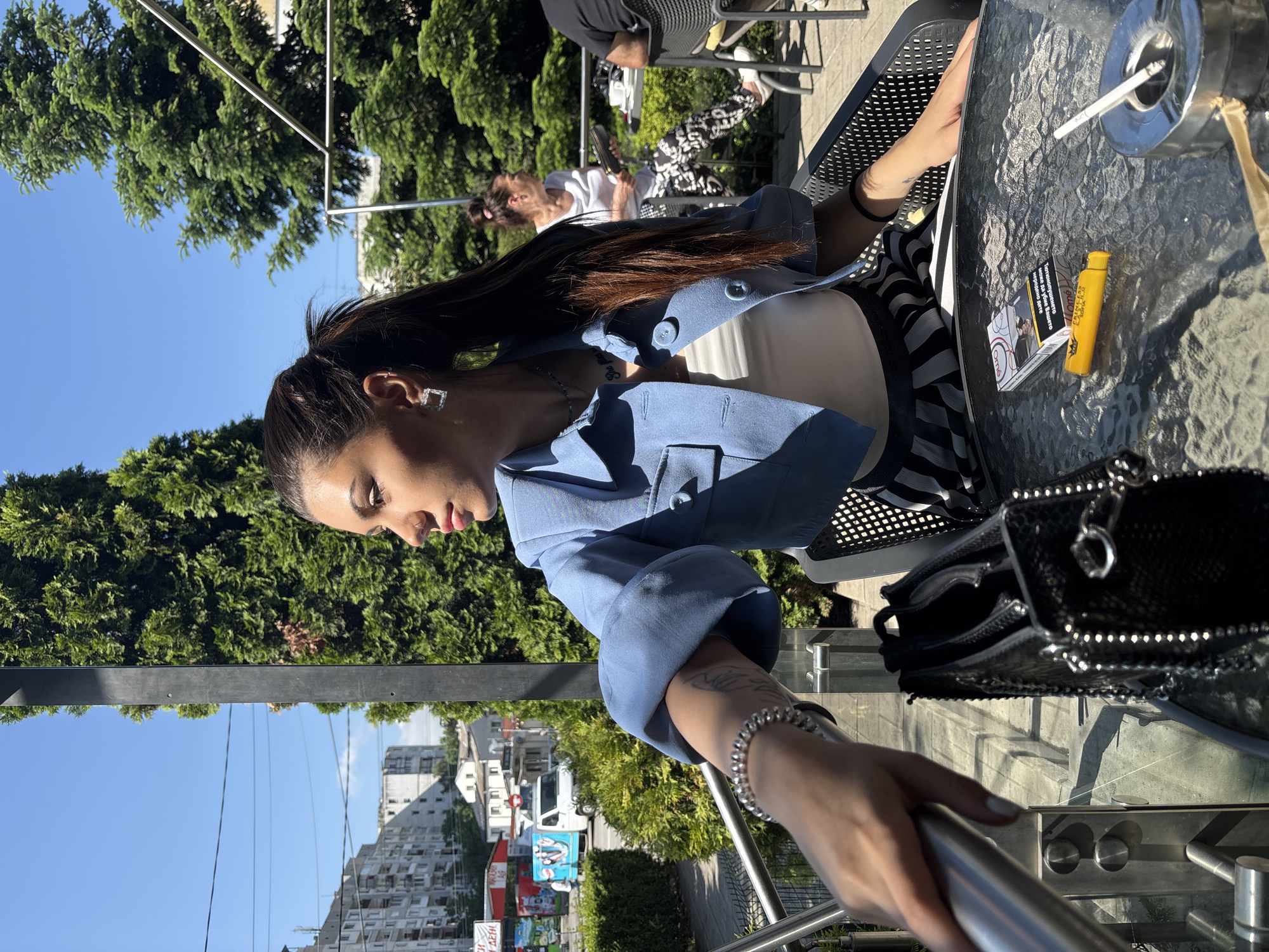 The image captures a casual outdoor scene at a café in Sofia, Bulgaria, on a sunny day. The main subject is a young woman with long dark hair styled in a high ponytail, seated at a clear glass table. She wears a light blue cropped jacket over a white top and a black and white striped skirt or trousers. Notable accessories include square silver earrings, a silver bracelet on her right wrist, and a visible tattoo on her right forearm. Her gaze is directed downwards, suggesting she might be looking at something on the table or lost in thought, creating a relaxed and contemplative mood.
On the glass table in front of her, a pack of cigarettes (partially revealing the brand "IQOS" and a health warning in Cyrillic, including the word "ПРЕДУПРЕЖДЕНИЕ" meaning "Warning"), a yellow lighter, and an ashtray with a cigarette resting in it are visible. A black, studded handbag is also on the table.
In the background, other patrons are seated at similar tables, including a woman with lighter hair wearing a white top and patterned pants. Lush green trees frame the upper and middle sections of the image, indicating a leafy or park-like urban environment. Beyond the trees, residential or commercial buildings are visible under a clear blue sky. A street scene with a white van and a colorful billboard is discernible further in the background. The bright sunlight casts distinct shadows, confirming it is daytime. The woman's hand is visible in the foreground, resting on a metal railing.