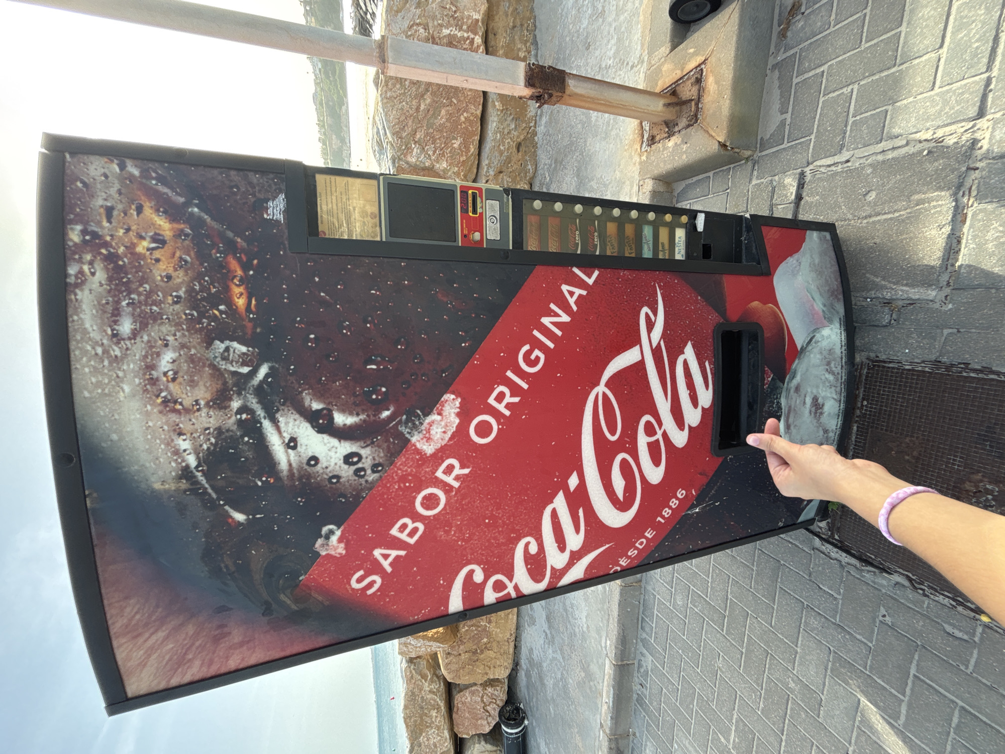 An outdoor Coca-Cola vending machine is positioned on a paved area near a rocky coastline. The machine displays a large "Sabor Original Coca-Cola" graphic. To the right of the machine, a rusty metal pole is visible, and beyond that, the ocean and a distant shoreline with buildings can be seen under a clear sky. In the foreground, a person's arm with a purple bracelet is extended towards the vending machine's dispensing slot, forming a heart shape with their fingers. The ground is paved with rectangular gray tiles. The scene appears to be in Urbanización Cabo Roig, Spain, indicated by the presence of a Coca-Cola vending machine in an outdoor, coastal setting consistent with the area.