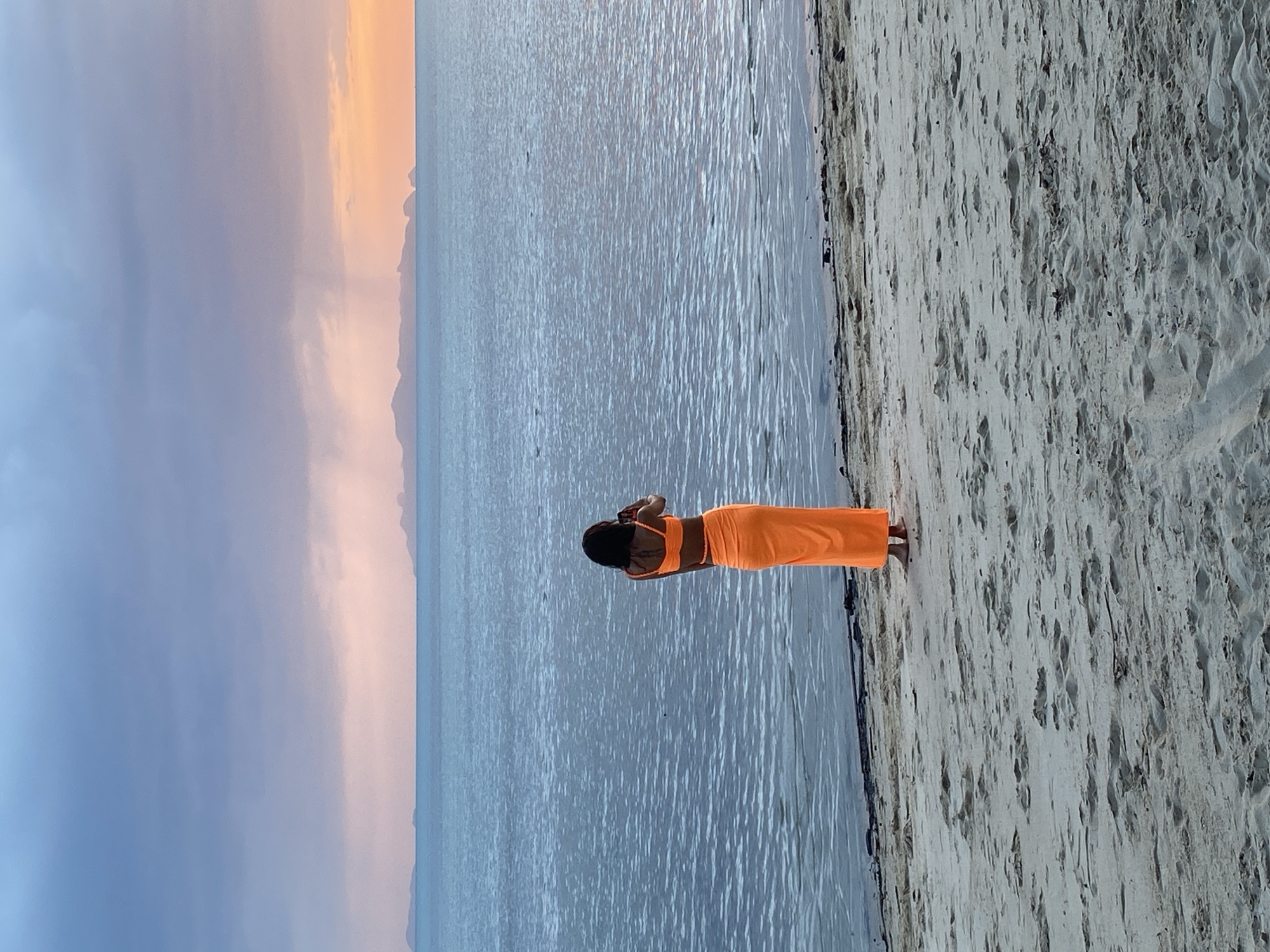 A person stands on a sandy beach, facing the ocean, in Ko Samui District, Thailand. The person, viewed from behind, wears an orange bikini top and a matching long skirt. They are holding a mobile phone to their ear, as if making a call or taking a picture. The ocean stretches out before them, with faint islands visible on the horizon. The sky above is a blend of blue, purple, and pink hues, suggesting either dawn or dusk. The sand is textured with footprints and debris.