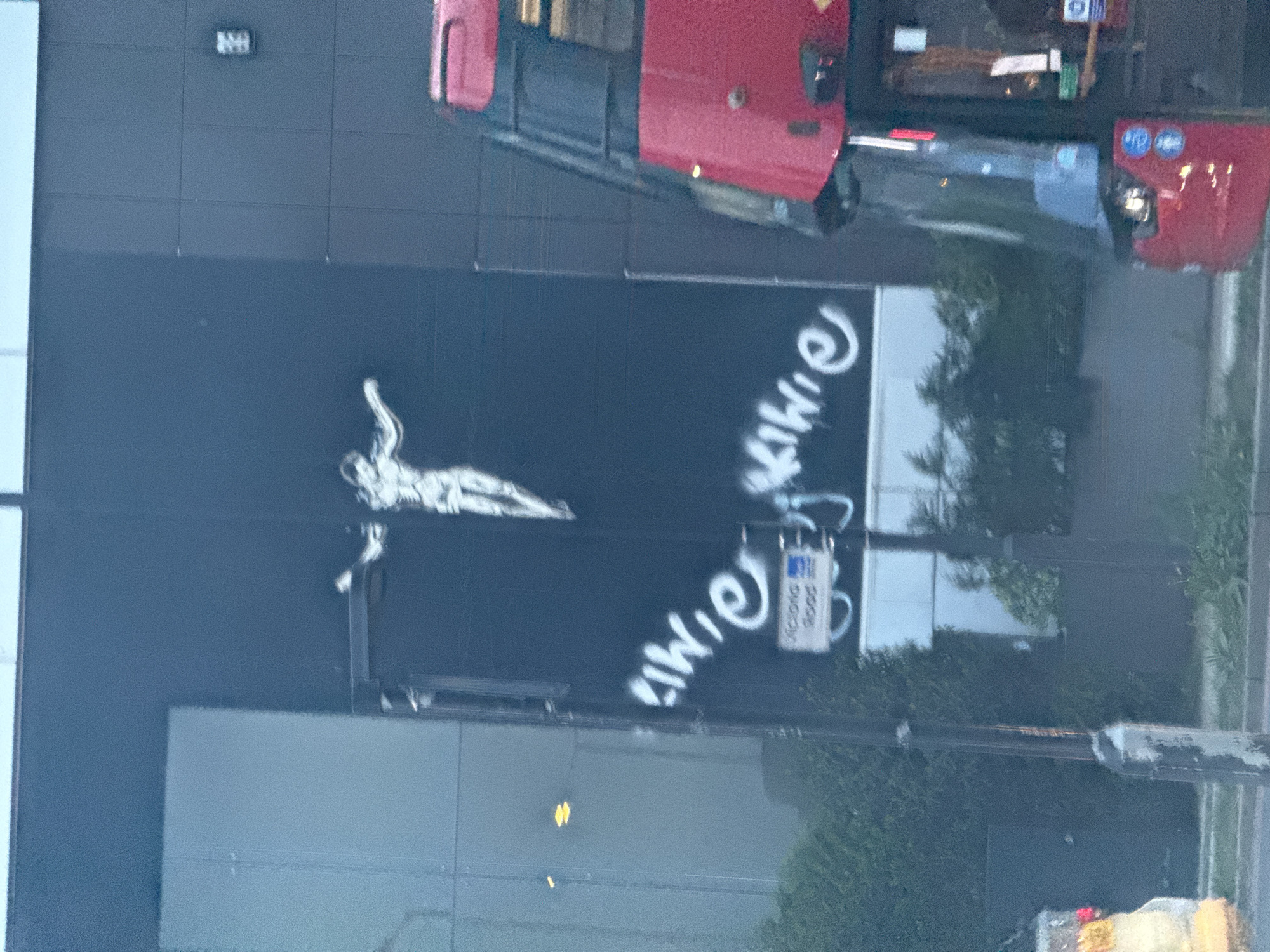 A street-level photograph taken in London, United Kingdom, shows the side of a building featuring a white painted figure in a reclining pose, along with the graffiti-style text "whiter shade". A red bus is visible in the upper portion of the image. A street sign is mounted on a pole in the foreground. The lighting suggests daylight.