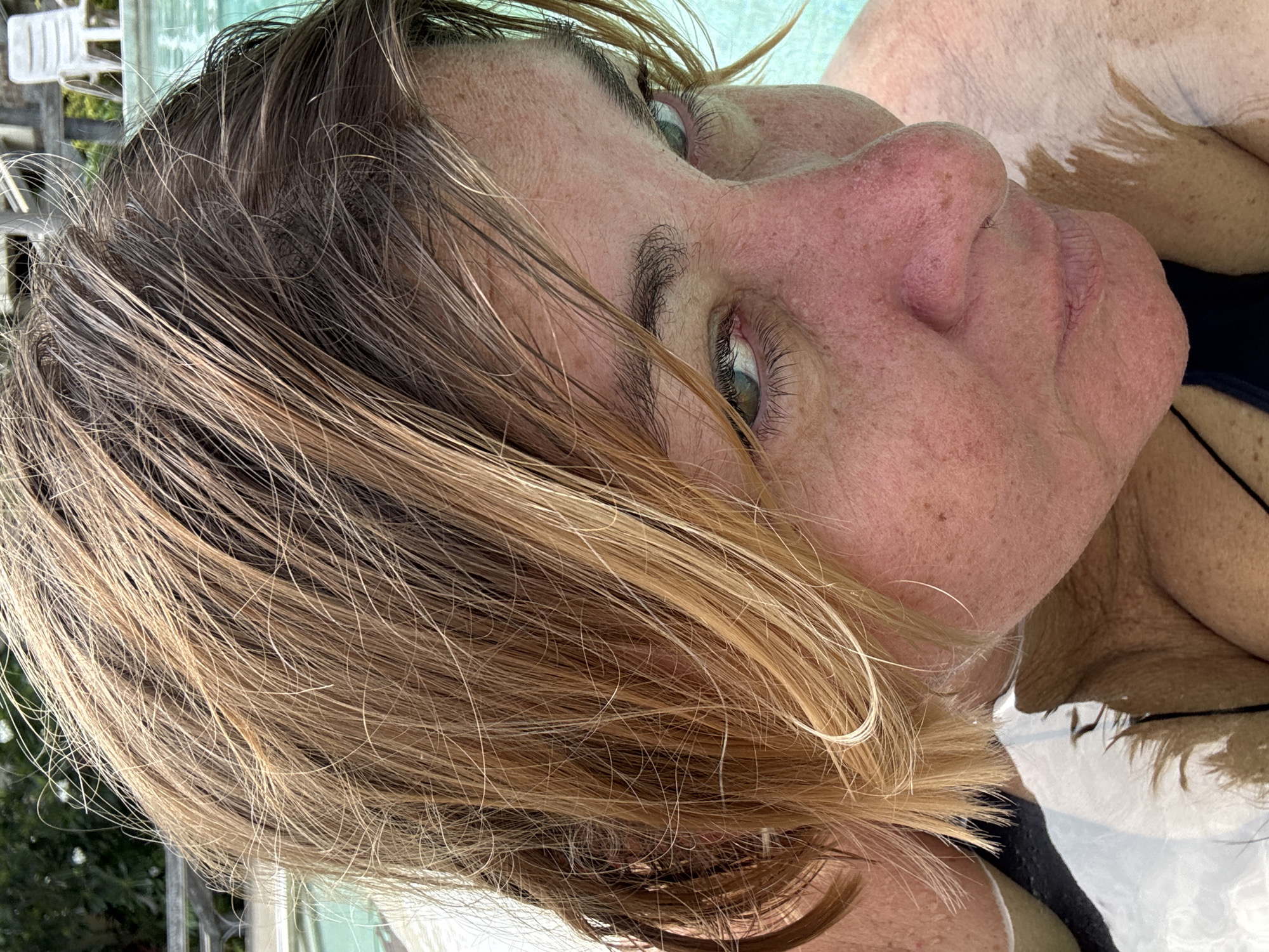 A close-up, rotated view of a person in water, likely a pool or tub. The person's face is angled towards the upper right, showcasing light skin with freckles and discoloration, particularly around the nose and eyes. The hair is wet, with light brown and blonde streaks. The individual appears to be submerged up to their neck and shoulders, with the water reflecting light. A black swimsuit strap is visible. The location is not discernible from this image.