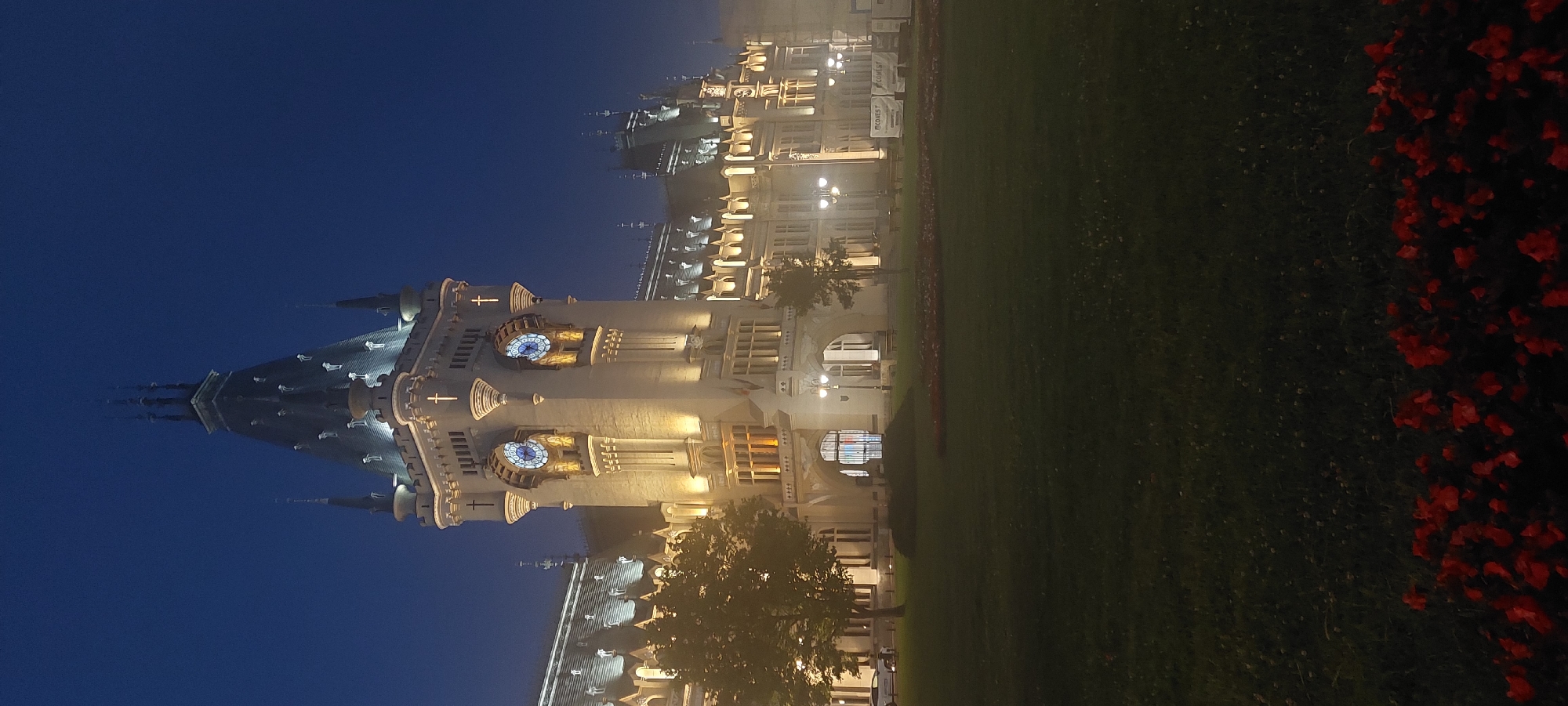 The Palace of Culture in Iași, Romania, is captured at twilight, with its imposing Gothic Revival architecture illuminated by warm artificial lights. The central tower, adorned with two large clock faces and intricate detailing, stands prominently against the deep blue of the evening sky. Tall, elegant windows and decorative crenellations line the extensive facade of the palace, which stretches horizontally across the frame.
In the foreground, a meticulously manicured lawn stretches out, bordered by a vibrant band of red flowers. The grass is slightly illuminated, revealing subtle textures and scattered small white flowers. To the left of the palace, mature trees with dark foliage stand as silhouettes against the building's glow.
Several lampposts, emitting a soft yellow light, are strategically placed along the pathways in front of the palace, highlighting architectural features and creating pools of light on the ground. In the middle distance, to the right of the main entrance, two white banners with text are visible. A few benches are also discernible in the same area. On the far left, a white car is parked near the edge of the lawn. The overall atmosphere is serene and majestic, enhanced by the atmospheric haze that slightly softens the distant details.